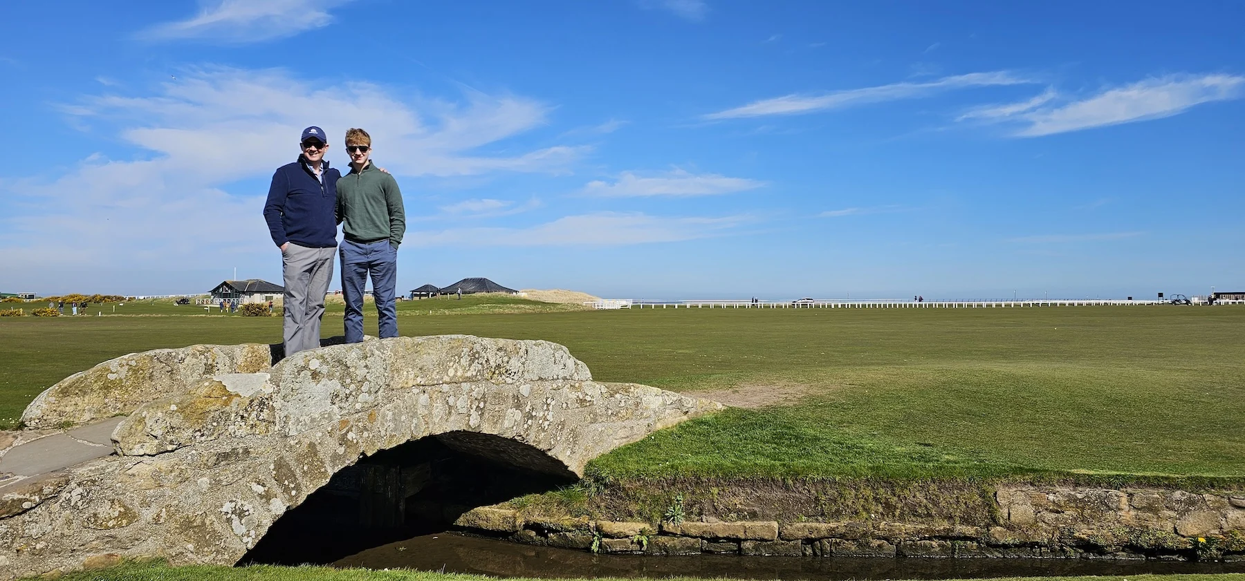 Two visitors stand on ancient stone bridge over Scottish countryside stream.