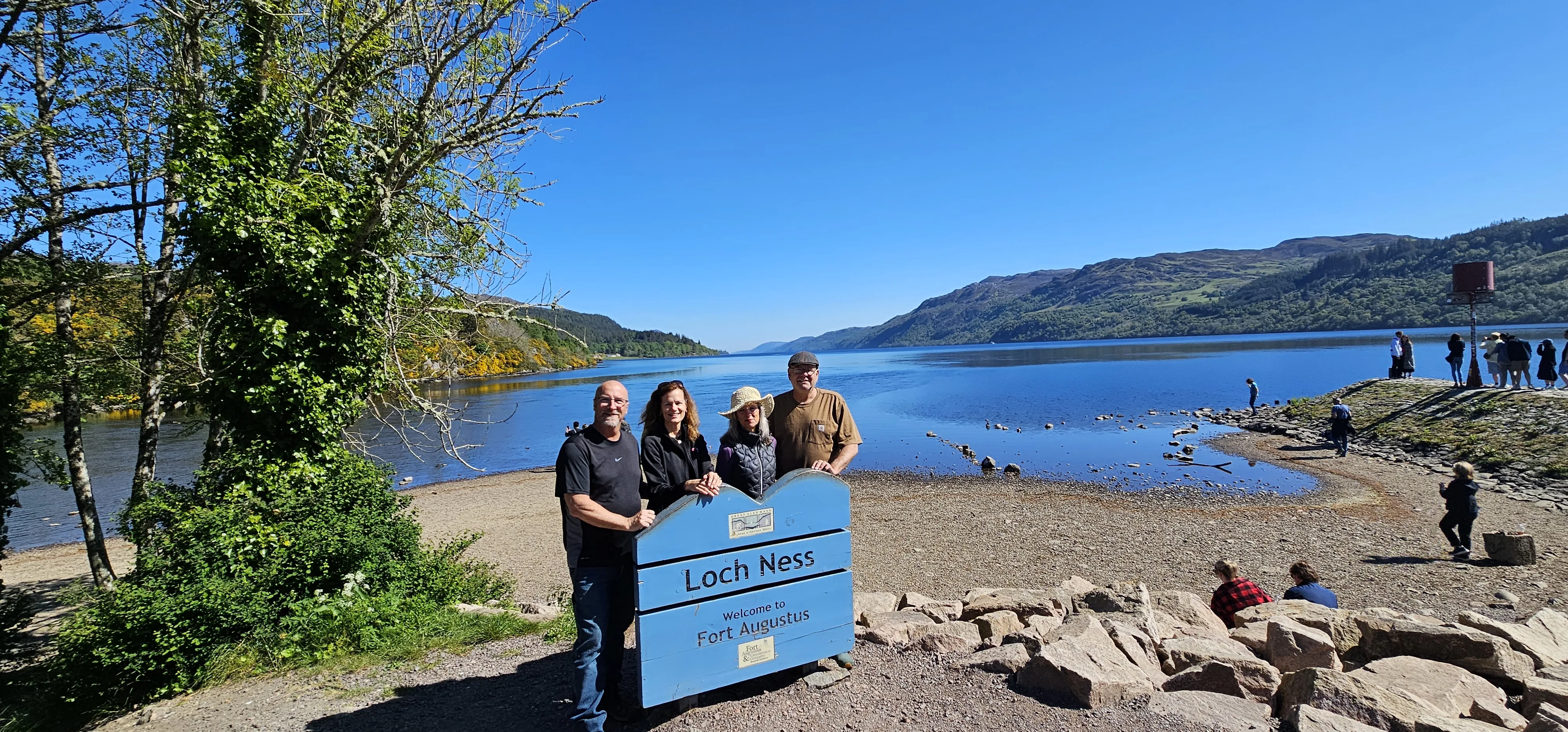Group posing at Loch Ness sign with scenic Highland landscape and water