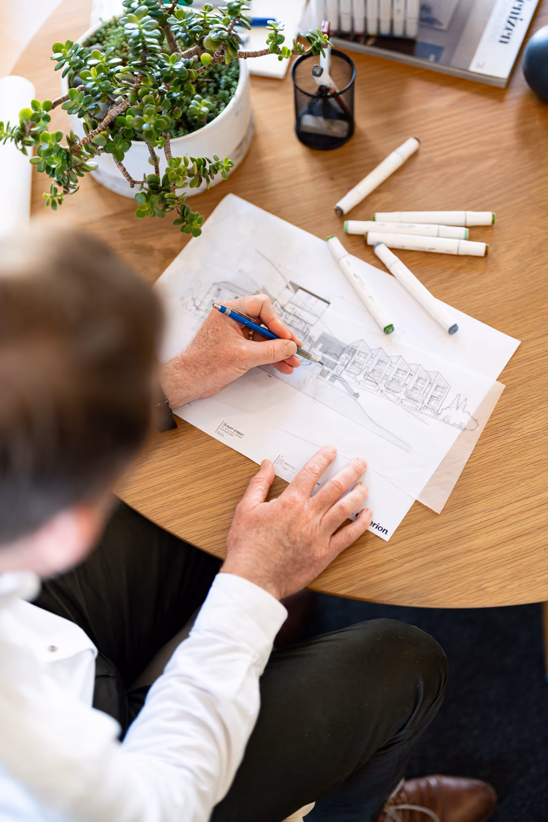 Person drawing architectural plans on paper at a wooden table with markers and a plant.