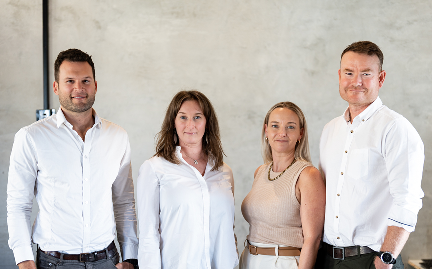 Four adults, two men and two women, standing side by side against a plain wall, smiling.