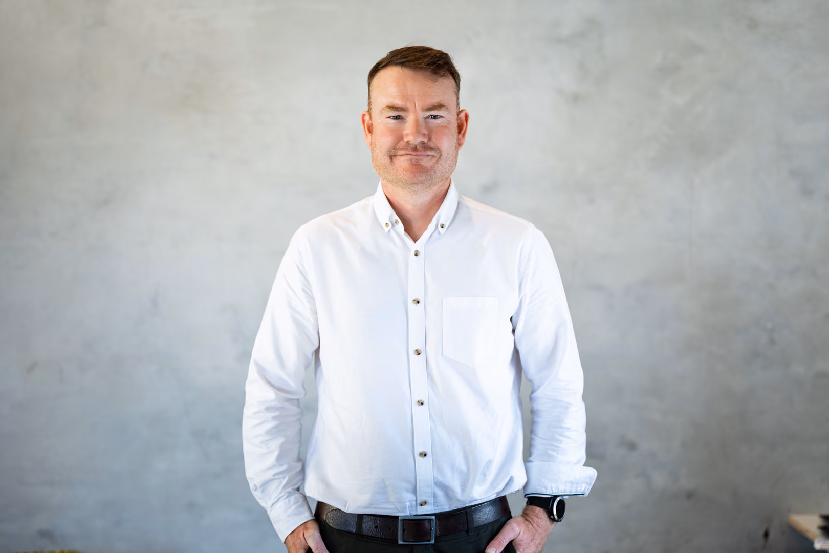 Smiling man with short hair, wearing a white button-up shirt and black belt, standing against gray wall.