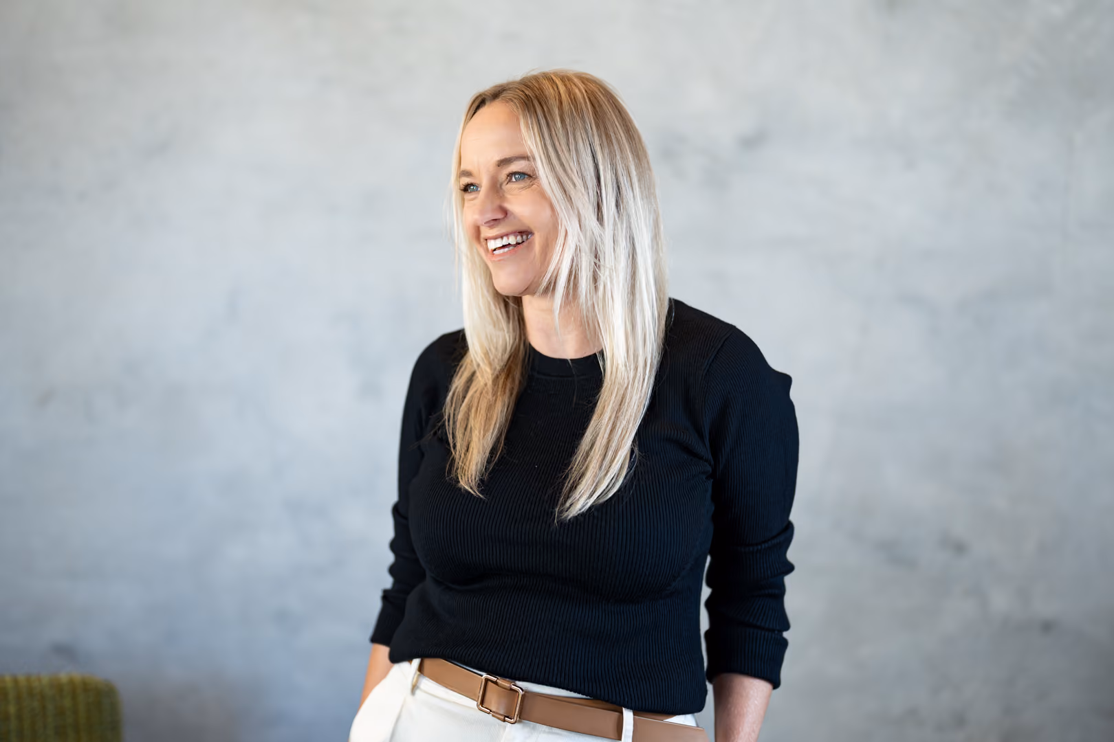 Smiling blonde woman in a black sweater and white pants stands against a gray textured wall.