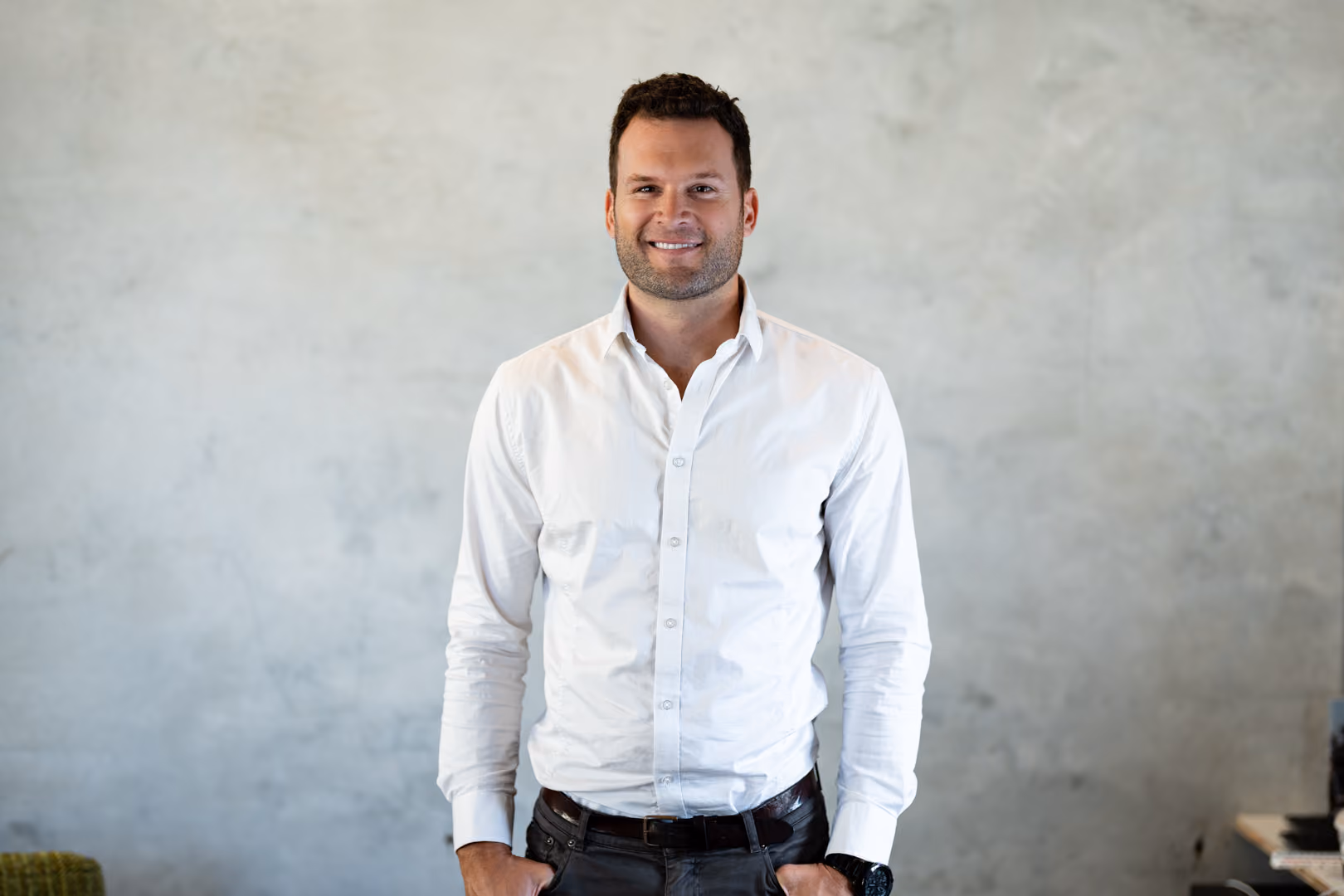 Smiling man wearing a white shirt and dark pants standing against a gray wall background.