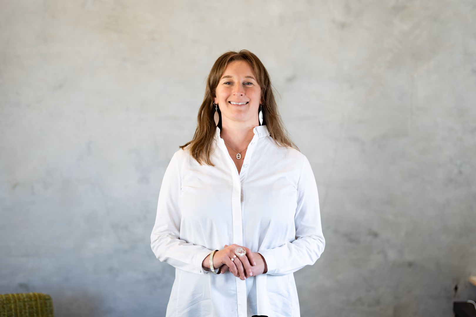 Smiling woman with brown hair wearing a white shirt stands against a plain gray wall.