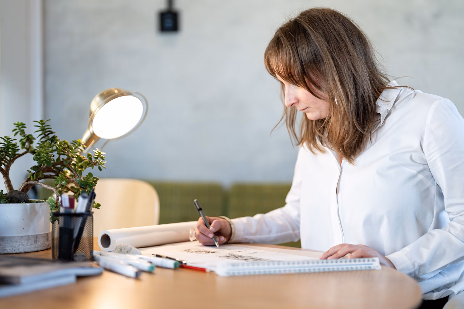 A woman in a white shirt sketches on a large pad at a desk with pens, a plant, and a lamp.