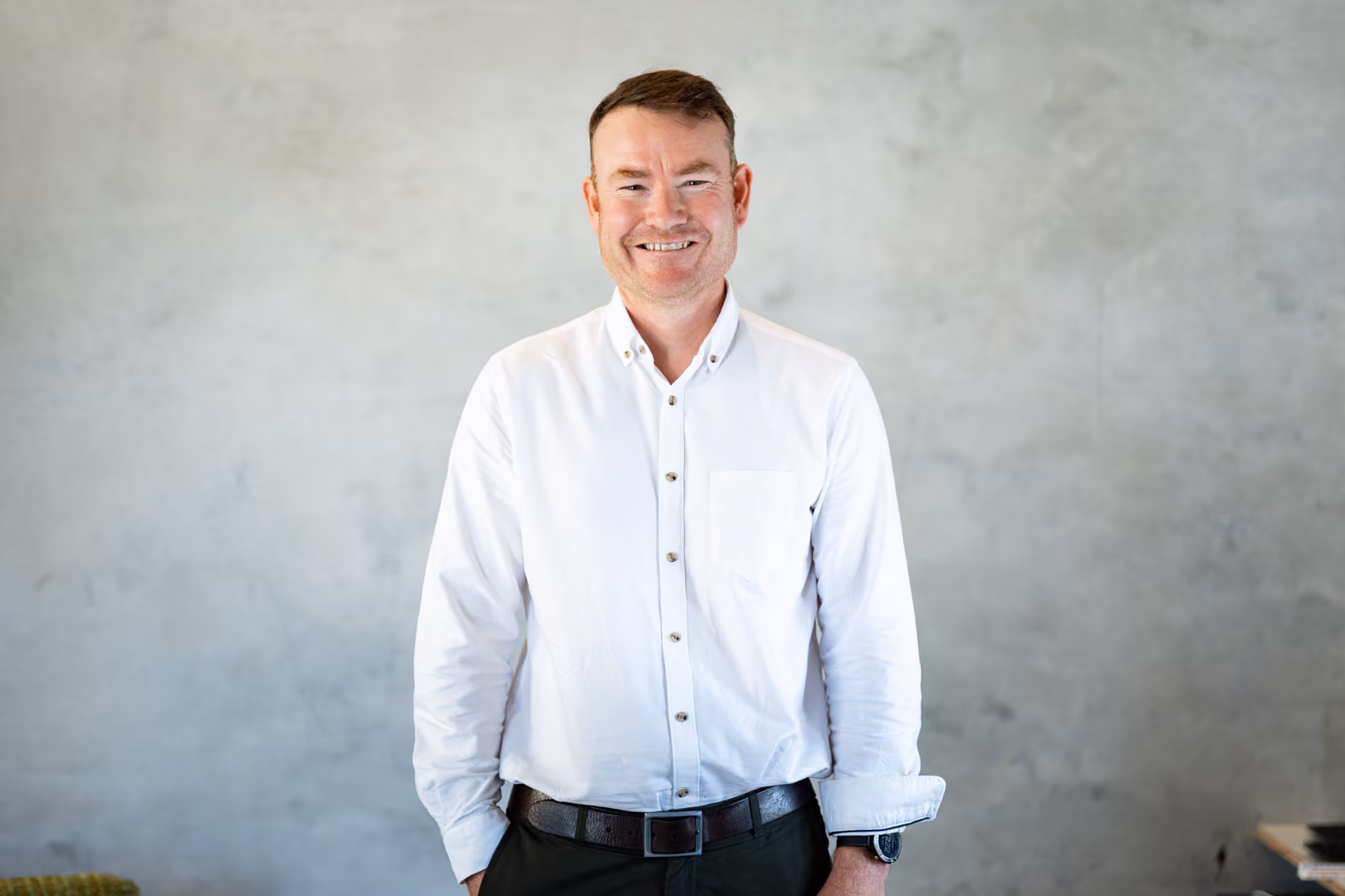Smiling man with short hair wearing a white button-up shirt and dark pants against a gray wall.