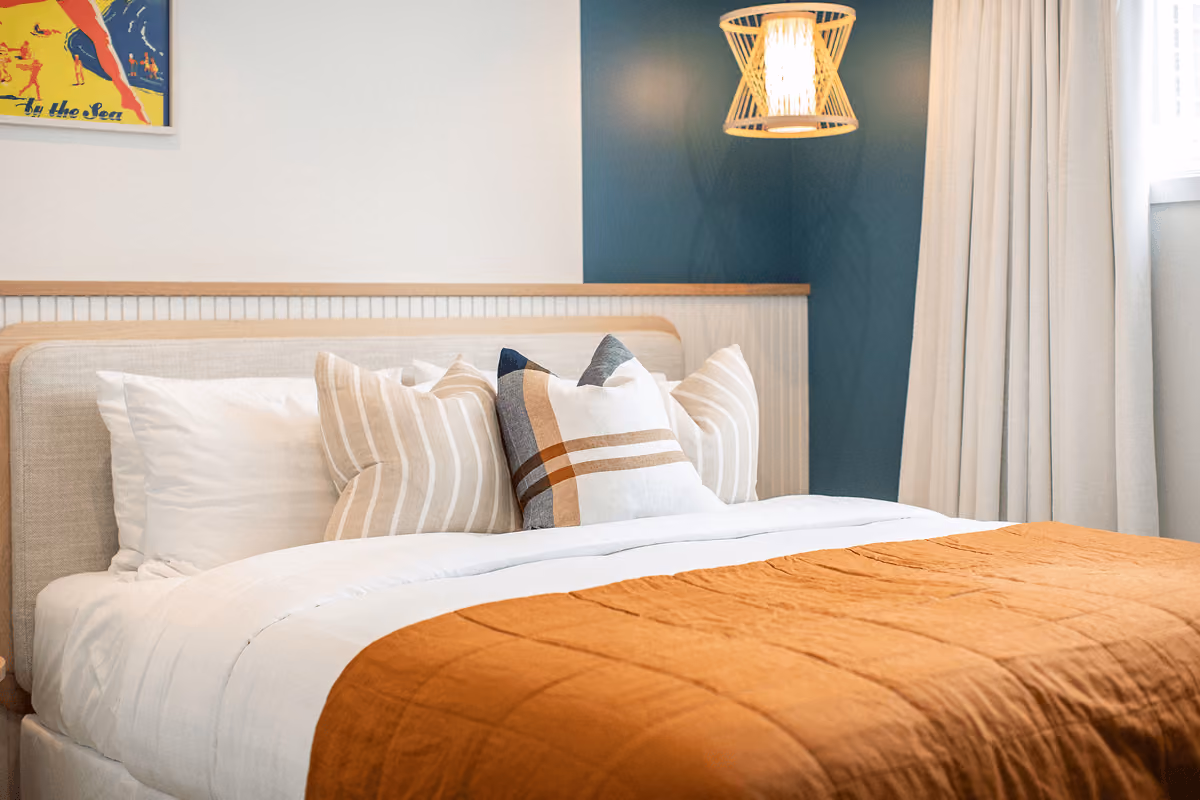 Bed with orange throw, patterned pillows, and a wooden lamp hanging on a teal accent wall.