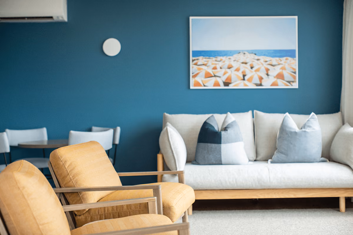 Living room with mustard armchairs, beige sofa with pillows, blue wall, and beach umbrella artwork.