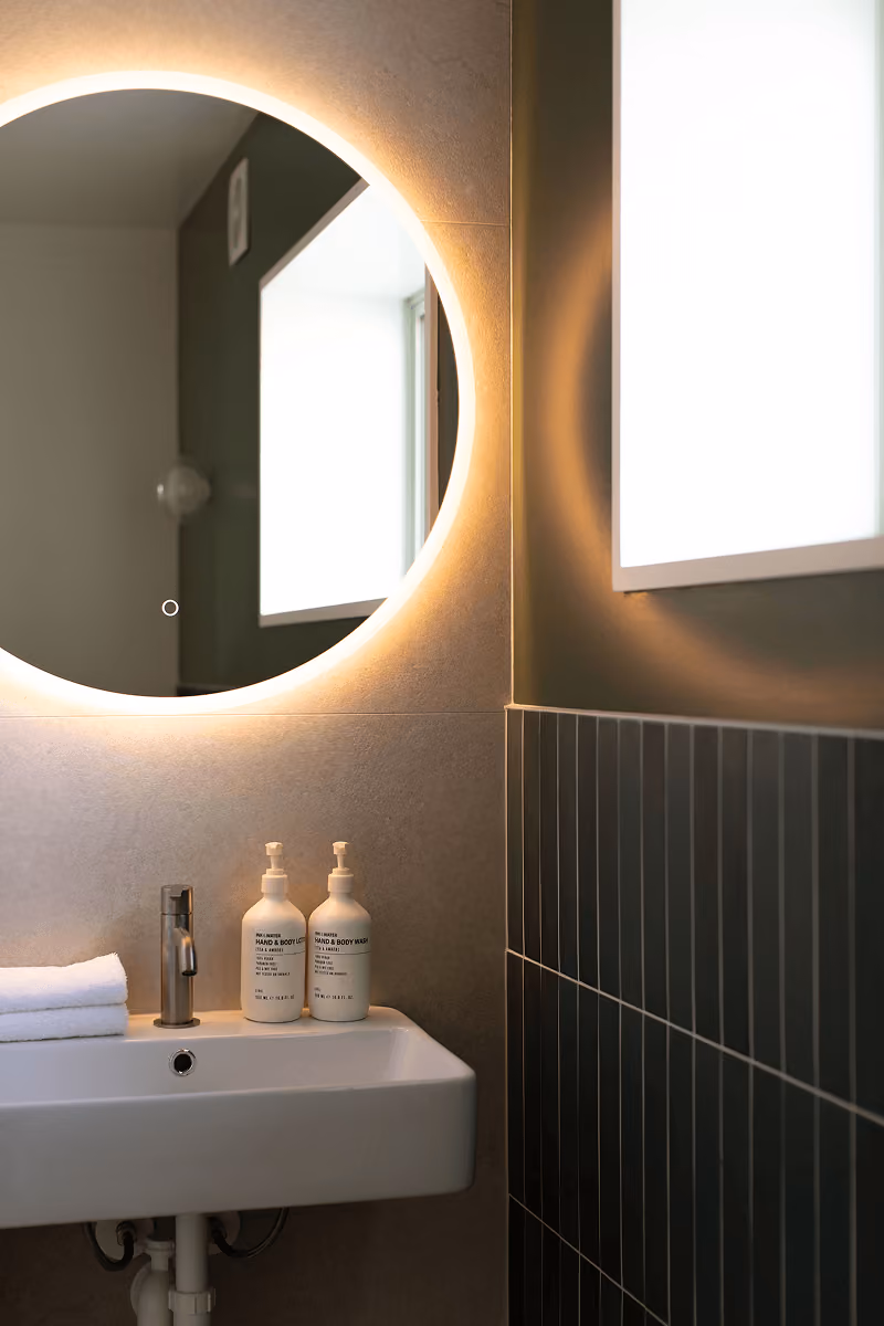 Modern bathroom sink with two hand and body wash bottles under a large round illuminated mirror.