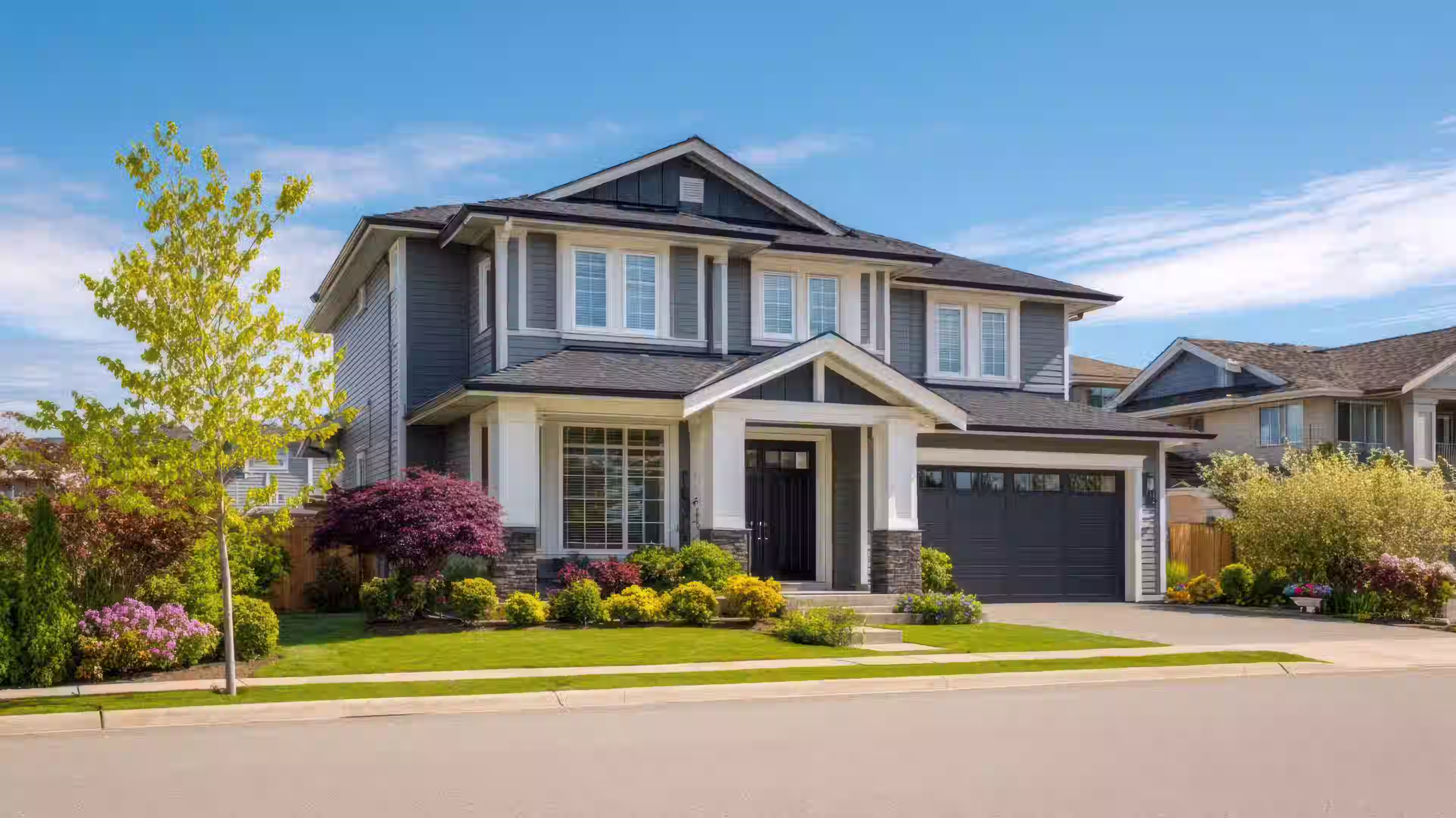 Two-story gray house with white trim, a black front door, garage, and landscaped yard with trees and shrubs.