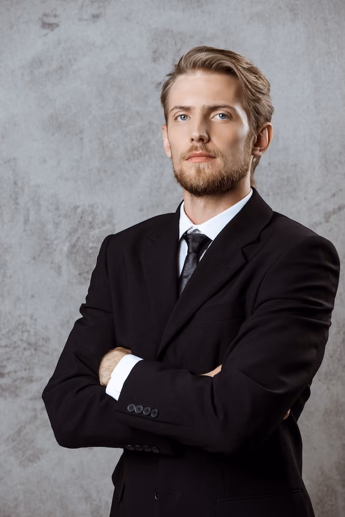 Portrait of a young man with beard in a black suit, white shirt, and black tie against a dark background.