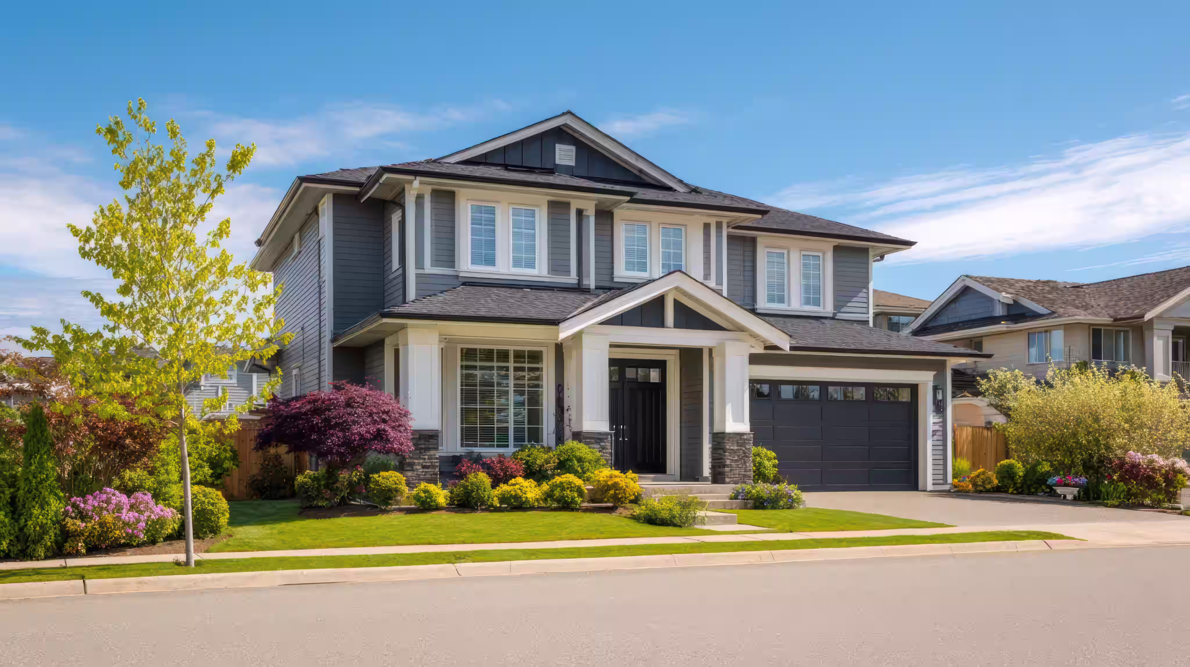 Modern two-story gray house with a black garage door, front porch, and colorful landscaping.
