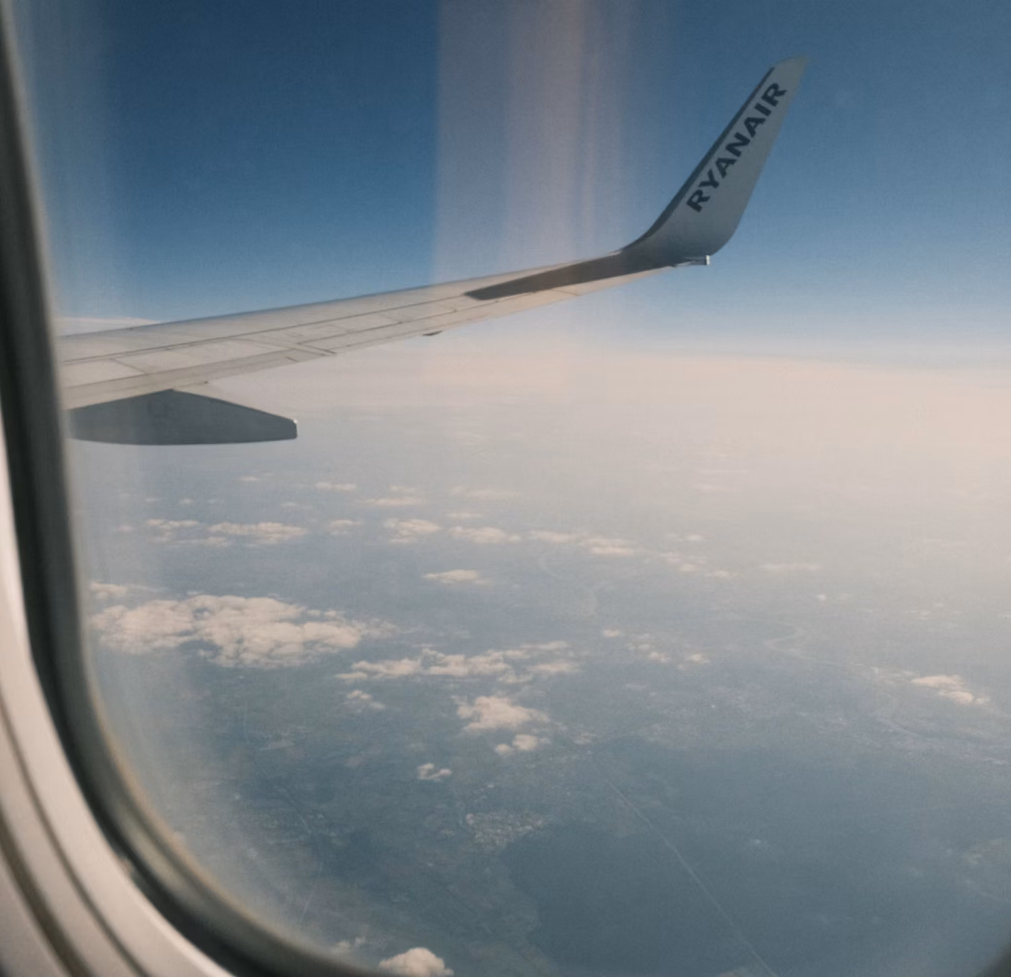 View from airplane window showing Ryanair wing and clouds over land beneath a clear sky.