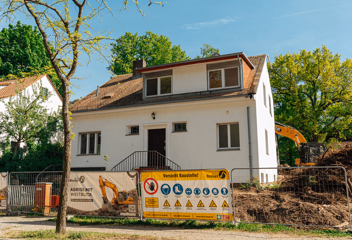 Weiße Einfamilienhaus vor Baustelle mit Bagger, Sicherheitszaun und Warnschildern, umgeben von Bäumen.