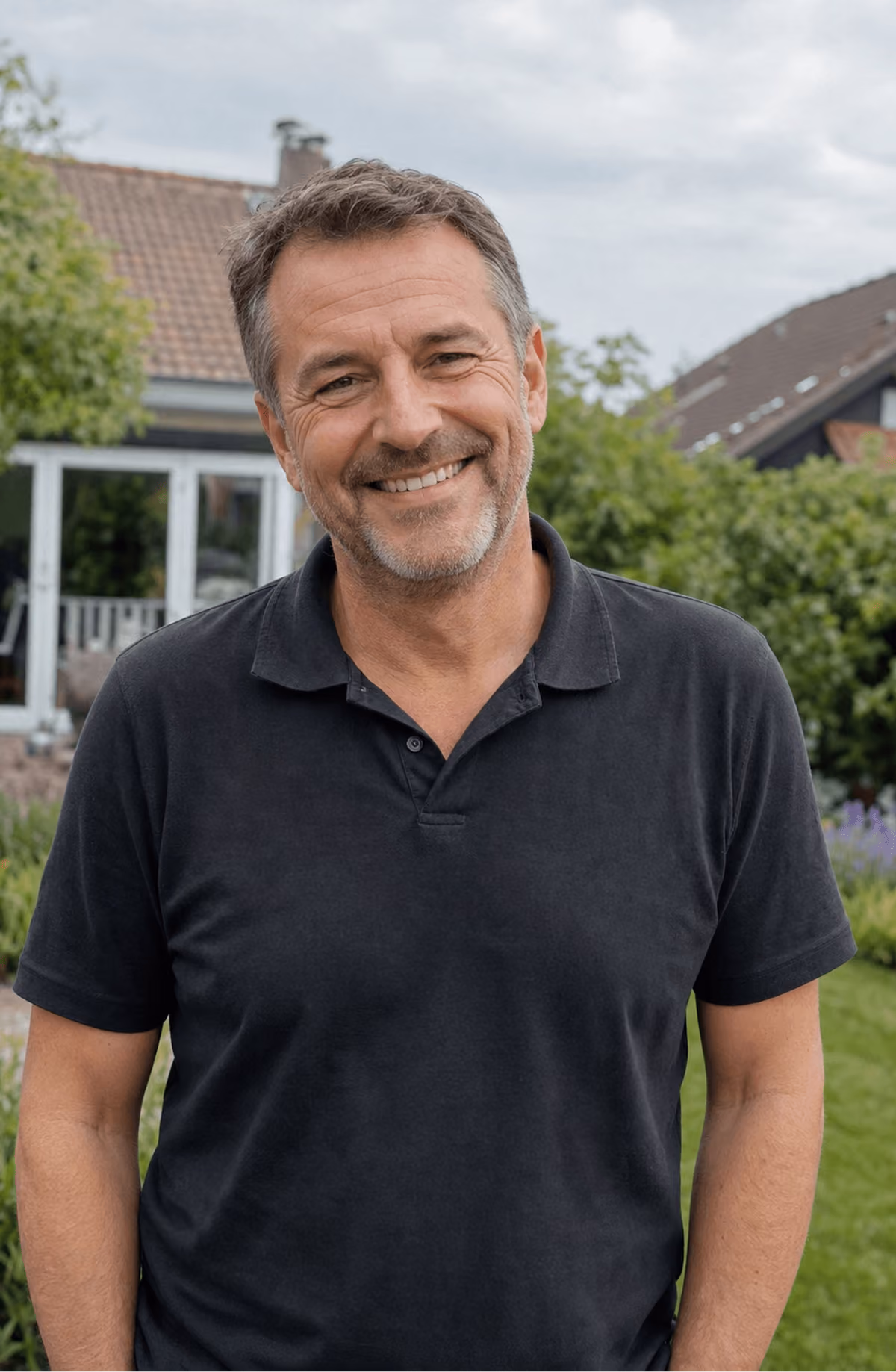 Mann mittleren Alters mit Bart lächelt im Garten; trägt schwarzes Polohemd; grüner Hintergrund mit Bäumen und Haus.