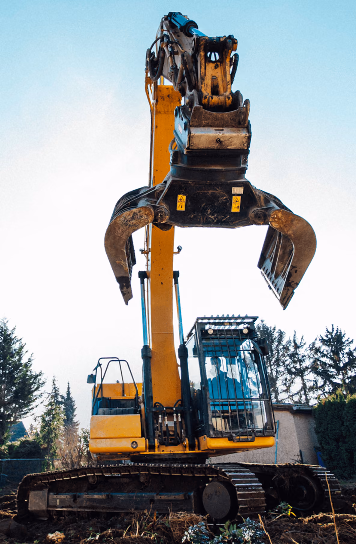 Gelber Bagger mit großem Greifarm im Einsatz auf Baustelle, umgeben von Bäumen und Himmel im Hintergrund.