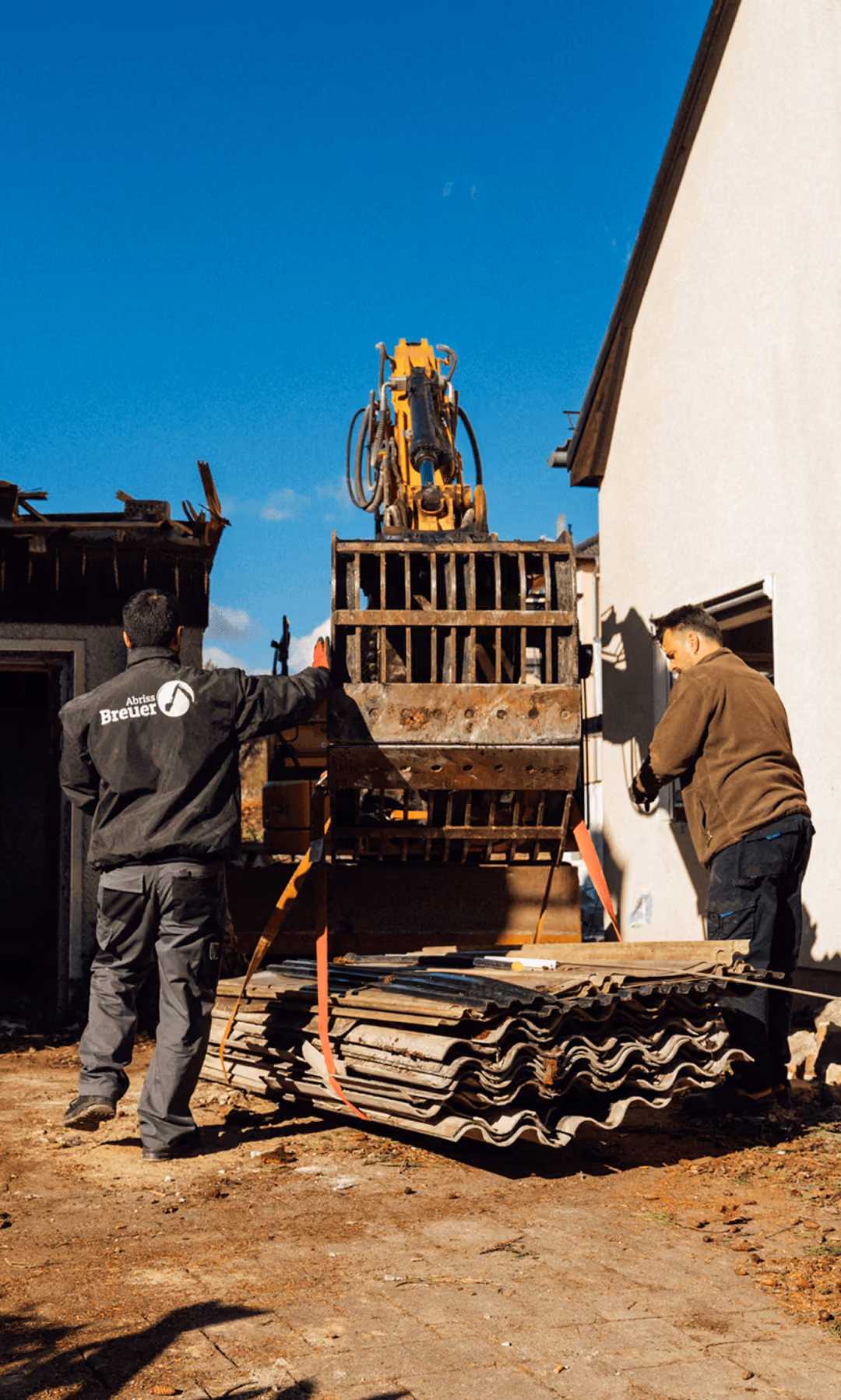 Bagger hebt Metallkäfig; zwei Männer sichern Last im Freien; blauer Himmel im Hintergrund.