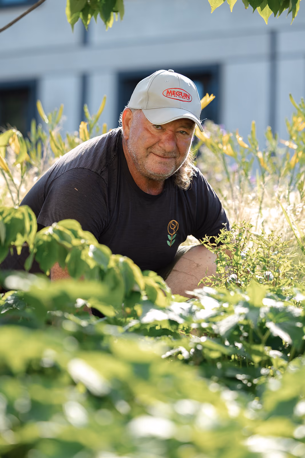 Ein älterer Mann mit grauem Bart und Cap kniet im Garten und lächelt, umgeben von grünen Pflanzen und Sonnenlicht.