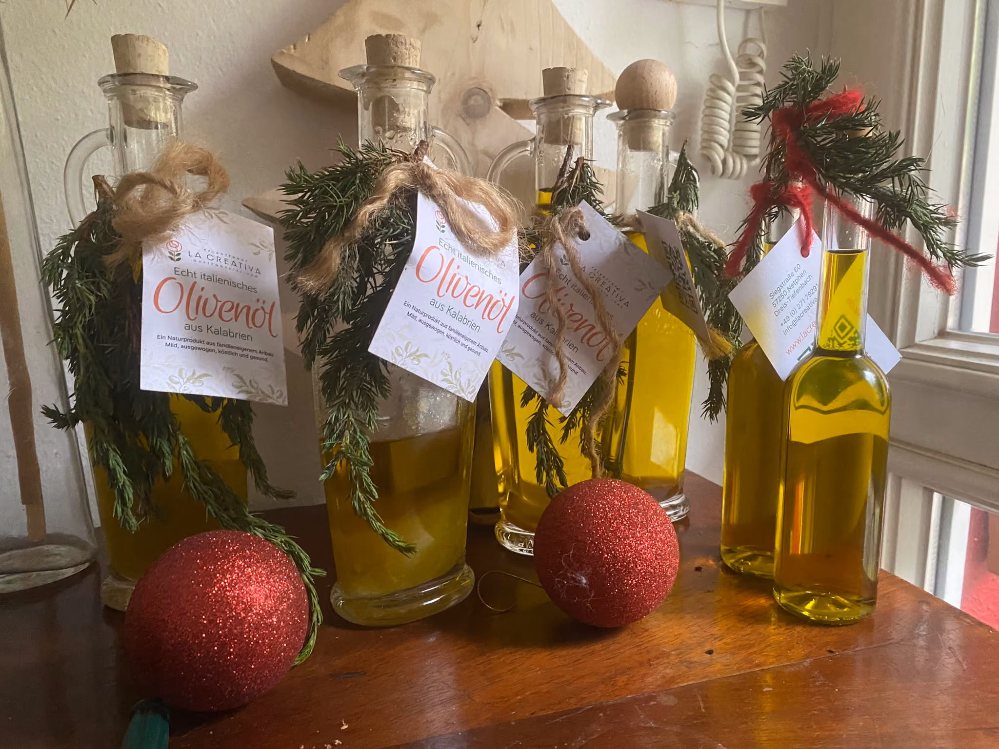 Fünf Glasflaschen mit echtem italienischem Olivenöl aus Kalabrien, dekoriert mit Korken, Zweigen Rosmarin und Etiketten, auf einem Holztisch neben zwei roten Weihnachtskugeln.
