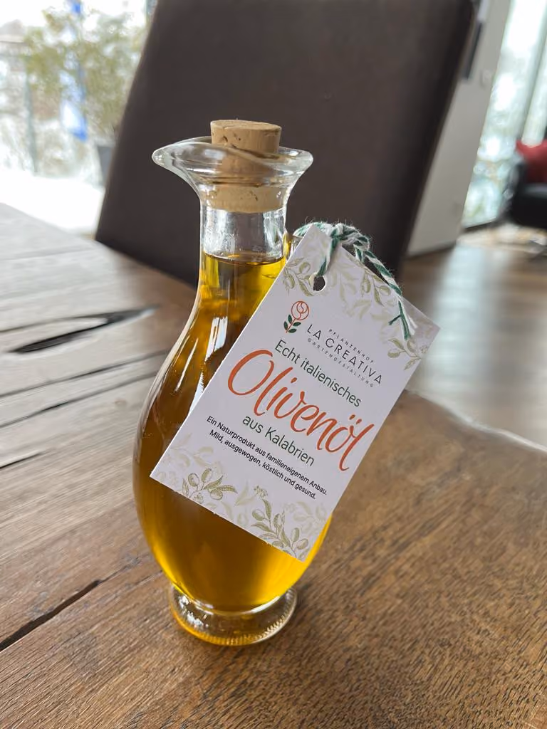 Eine Glasflasche mit Korken, gefüllt mit goldenem Olivenöl, steht auf einem Holztisch. An der Flasche hängt ein Etikett mit der Aufschrift 'Echt italienisches Olivenöl aus Kalabrien' und einer Beschreibung, dass es ein naturreines Produkt aus familieneigenem Anbau ist.