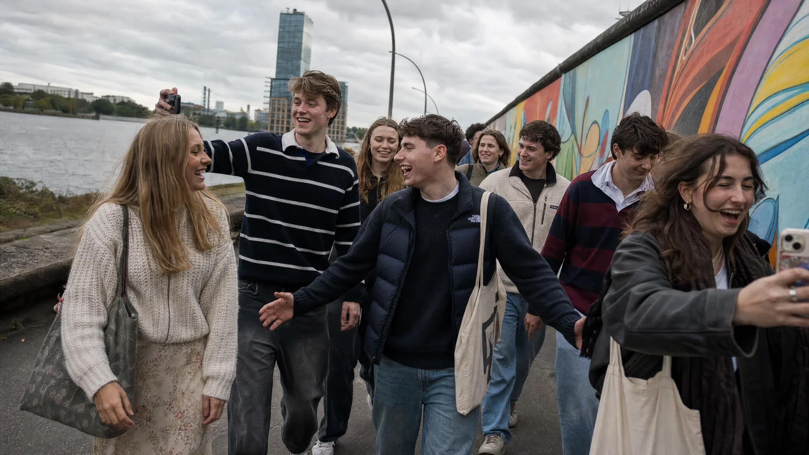 Group of young travellers laughing and taking selfies beside the East Side Gallery murals in Berlin.