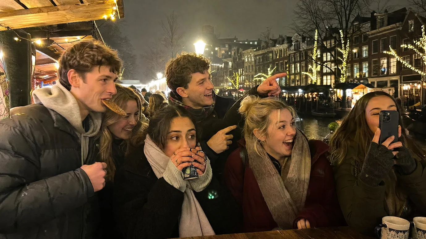 Group of young travellers enjoying warm drinks at an Amsterdam Christmas market with festive canal lights behind.
