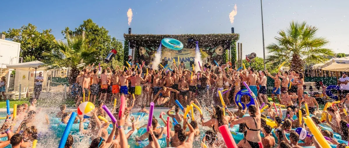 Crowd of partygoers cheering with pool floats and inflatables at a lively outdoor pool party with flame jets.