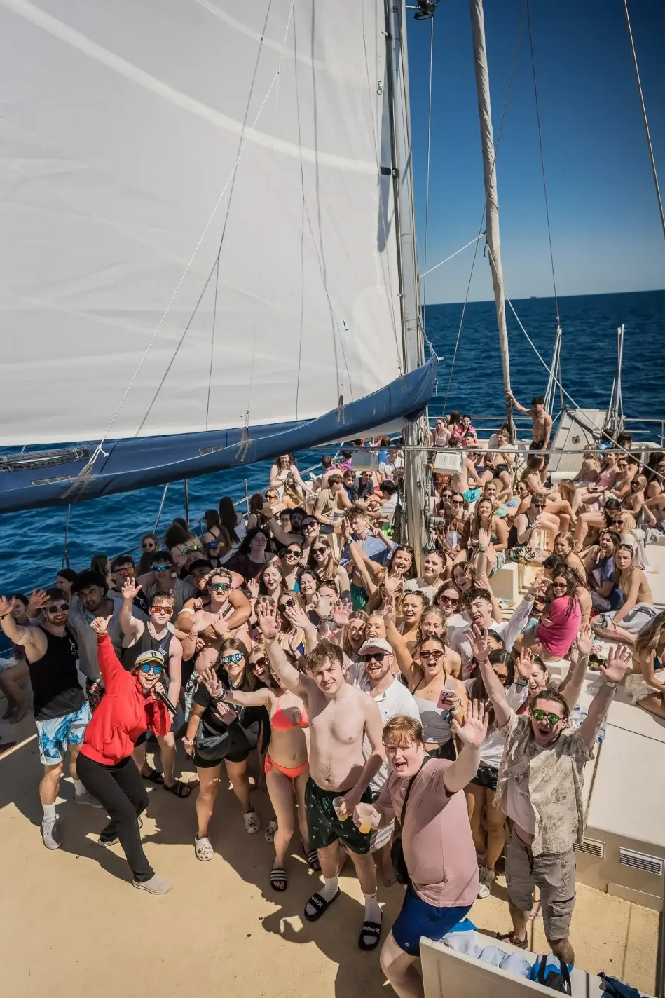 Group of young travellers waving from the deck of a sailing boat party at sea under bright sun.