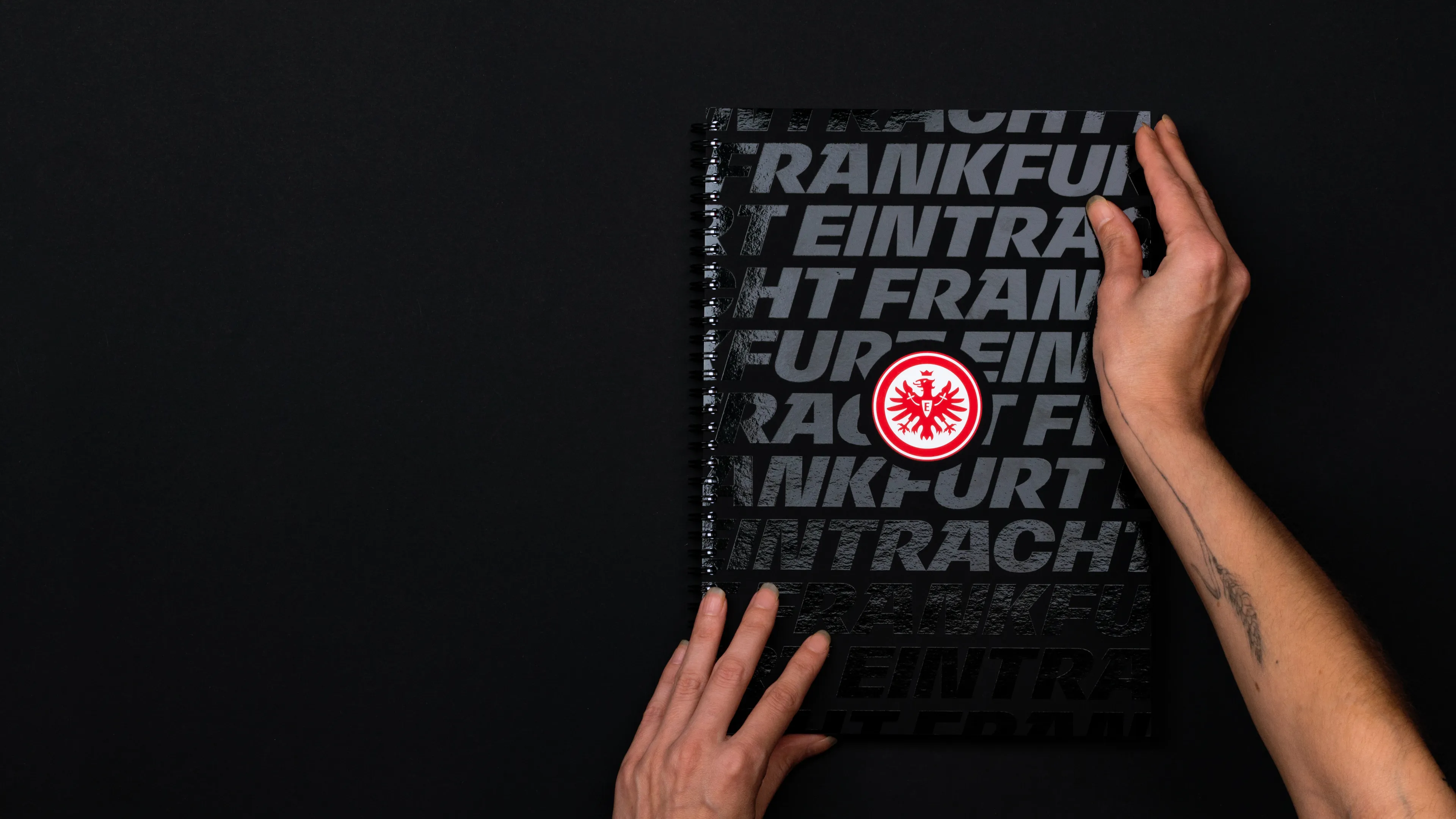 Eintracht Frankfurt Brandbook Cover. Zu sehen ist das Cover des Eintracht Frankfurt Brandbooks. Zwei Hände halten das Buch. Das schwarze Cover ist mit einer schwarzen Folienkaschierung veredelt. Die Veredelung zeigt den Eintracht Frankfurt Schriftzug. In der Mitte ist das Wappen des Vereins zu sehen.