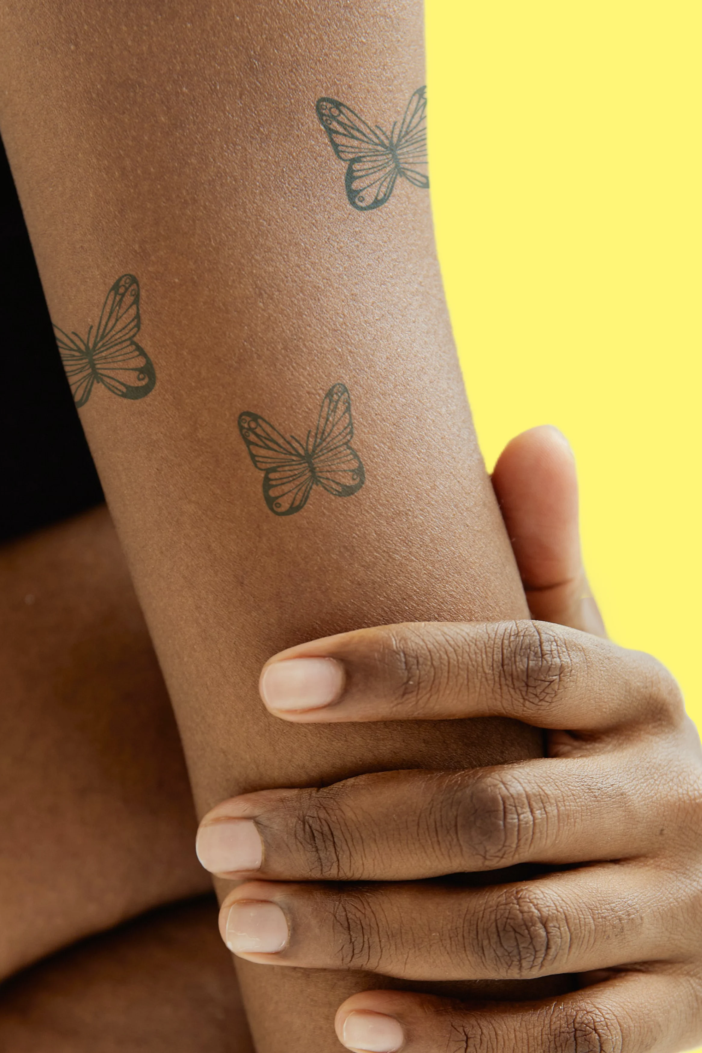 Nahaufnahme einer Person, die ihren Arm mit der anderen Hand umfasst. Auf dem Oberarm sind drei Schmetterlings-Tattoos zu erkennen.