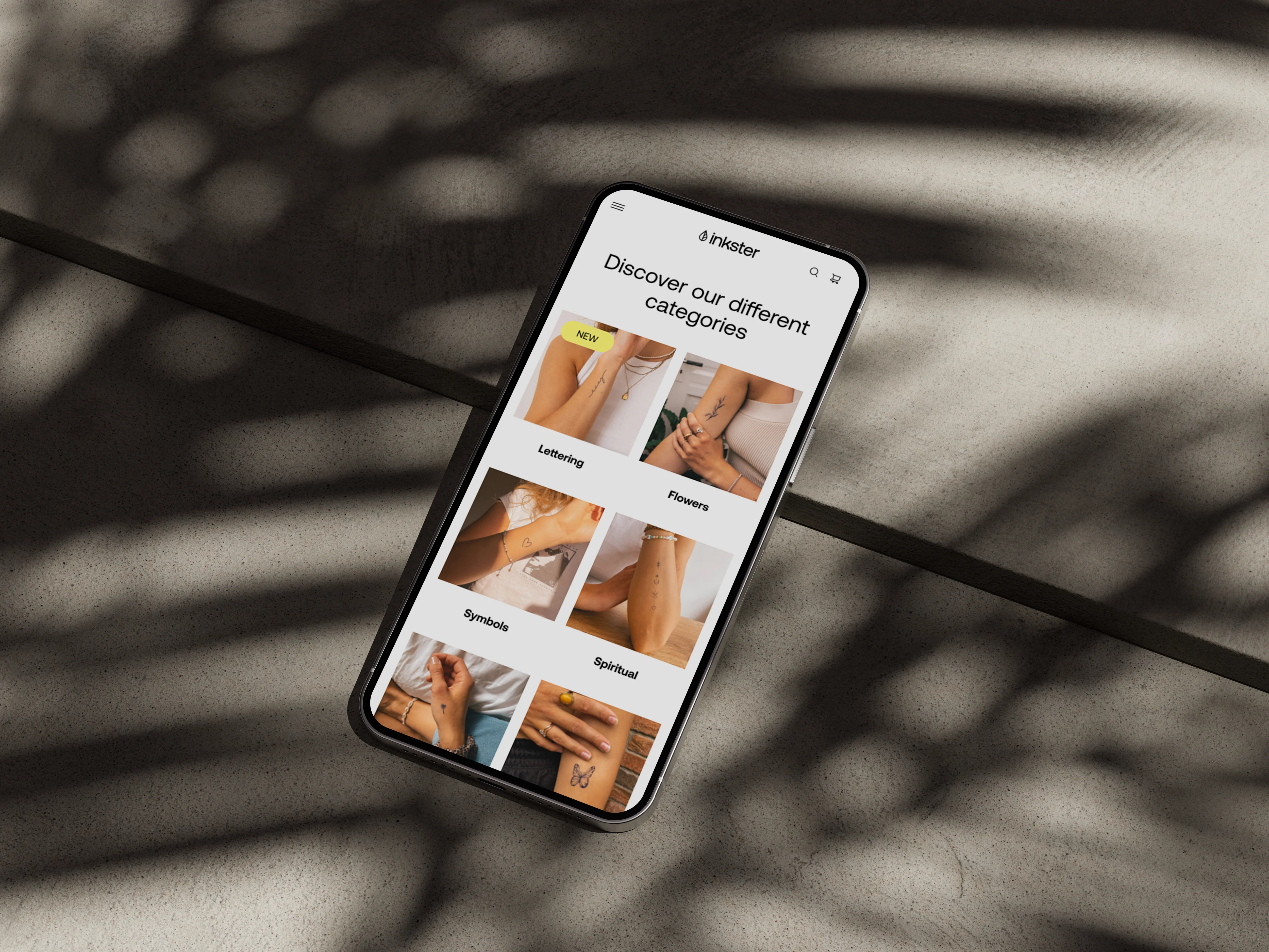 Ein iPhone liegt auf einem Steinboden, der Schatten einer Palme liegt quer über dem Bild. Der Bildschirm zeigt verschiedene Stilkategorien von Inkster-Tattoos.