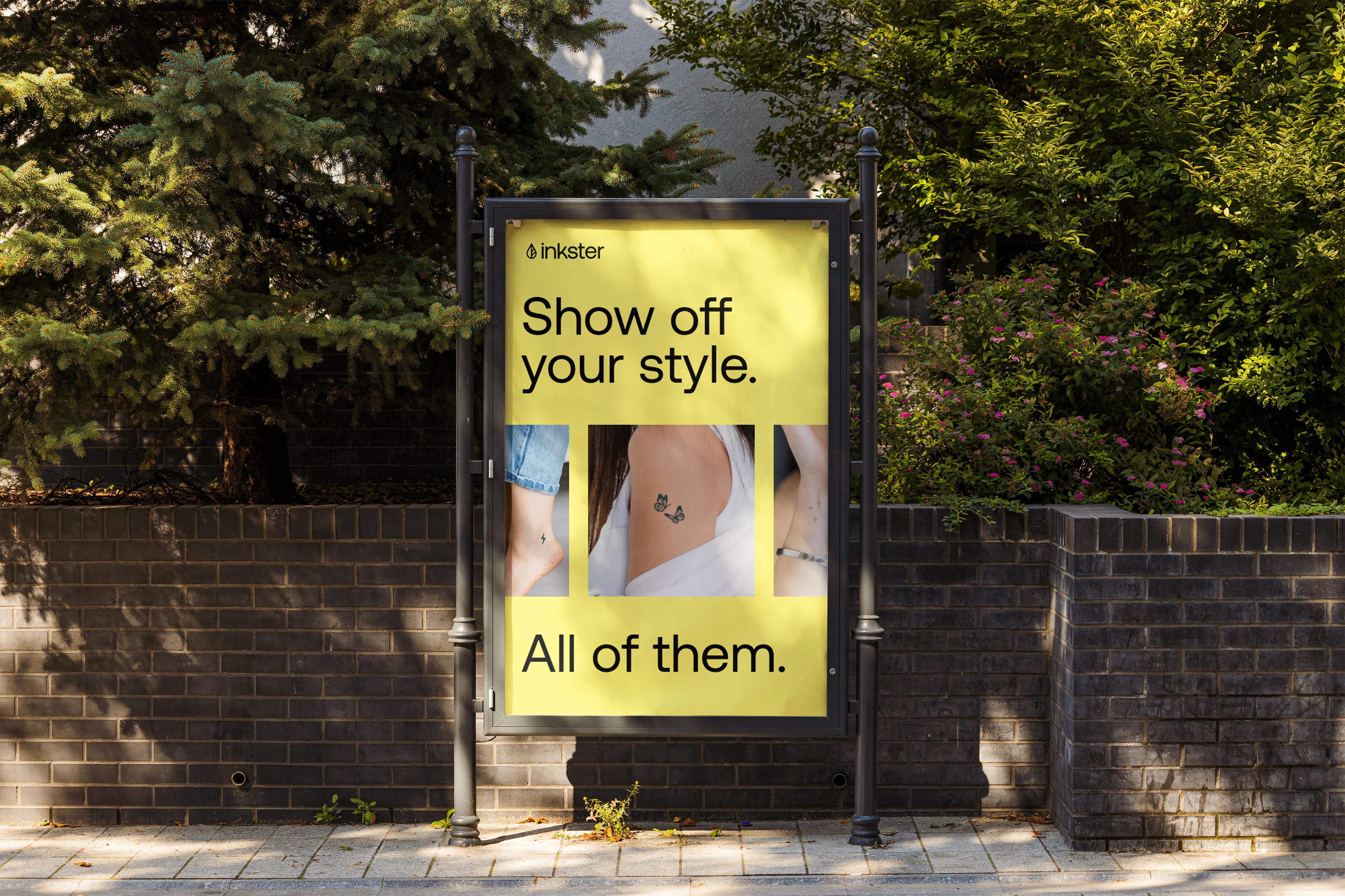 Ein Plakat hängt in einer sonnigen Straße mit Bäumen hinter einer Mauer. Das Plakat zeigt verschiedene Tattoos an verschiedenen Körperstellen und der Text lautet: "Show off your Style. All of them."