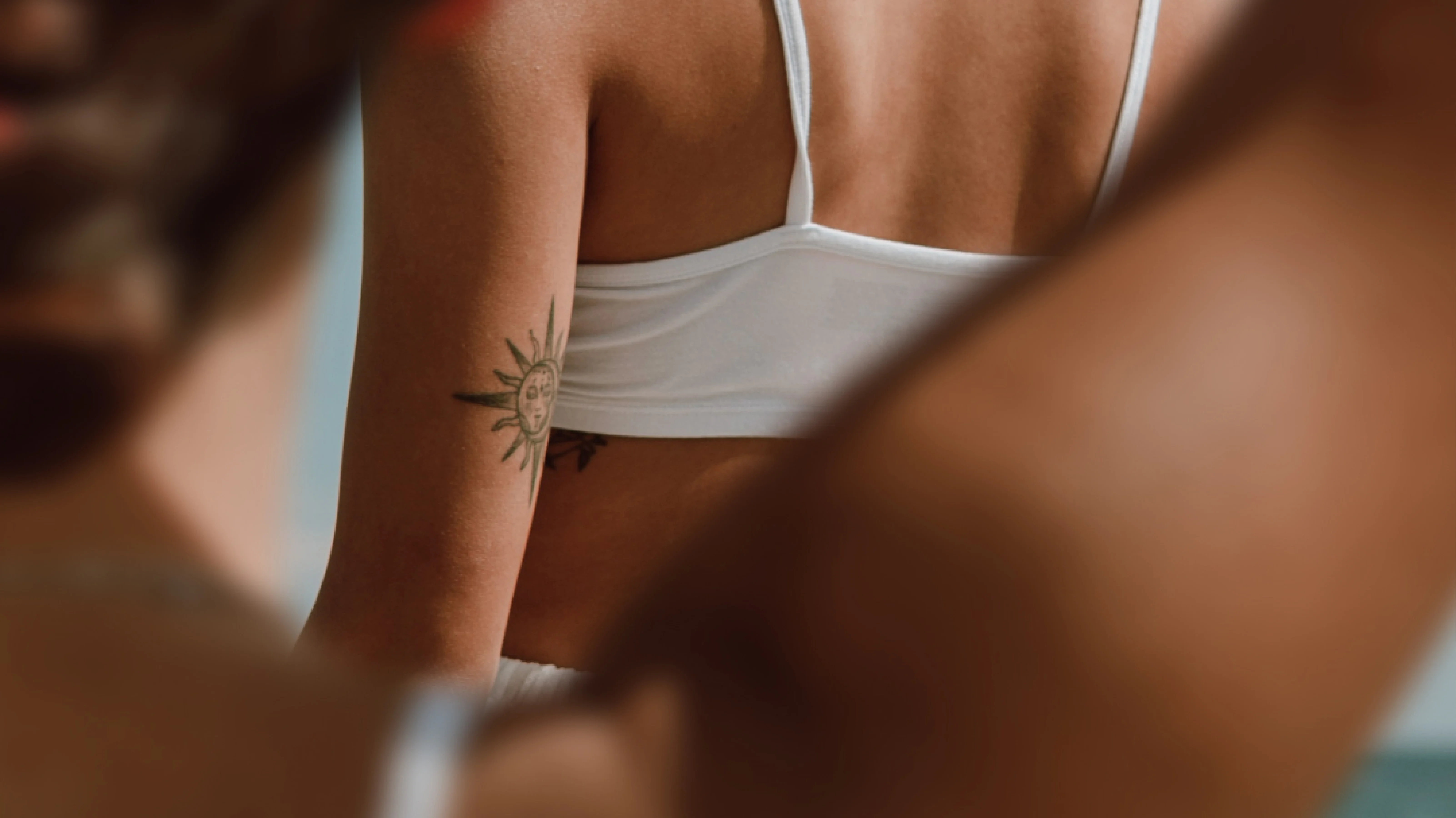 Eine Nahaufnahme zweier Personen. Die hintere Person trägt ein Sonnen-Tattoo auf ihrem Oberarm.