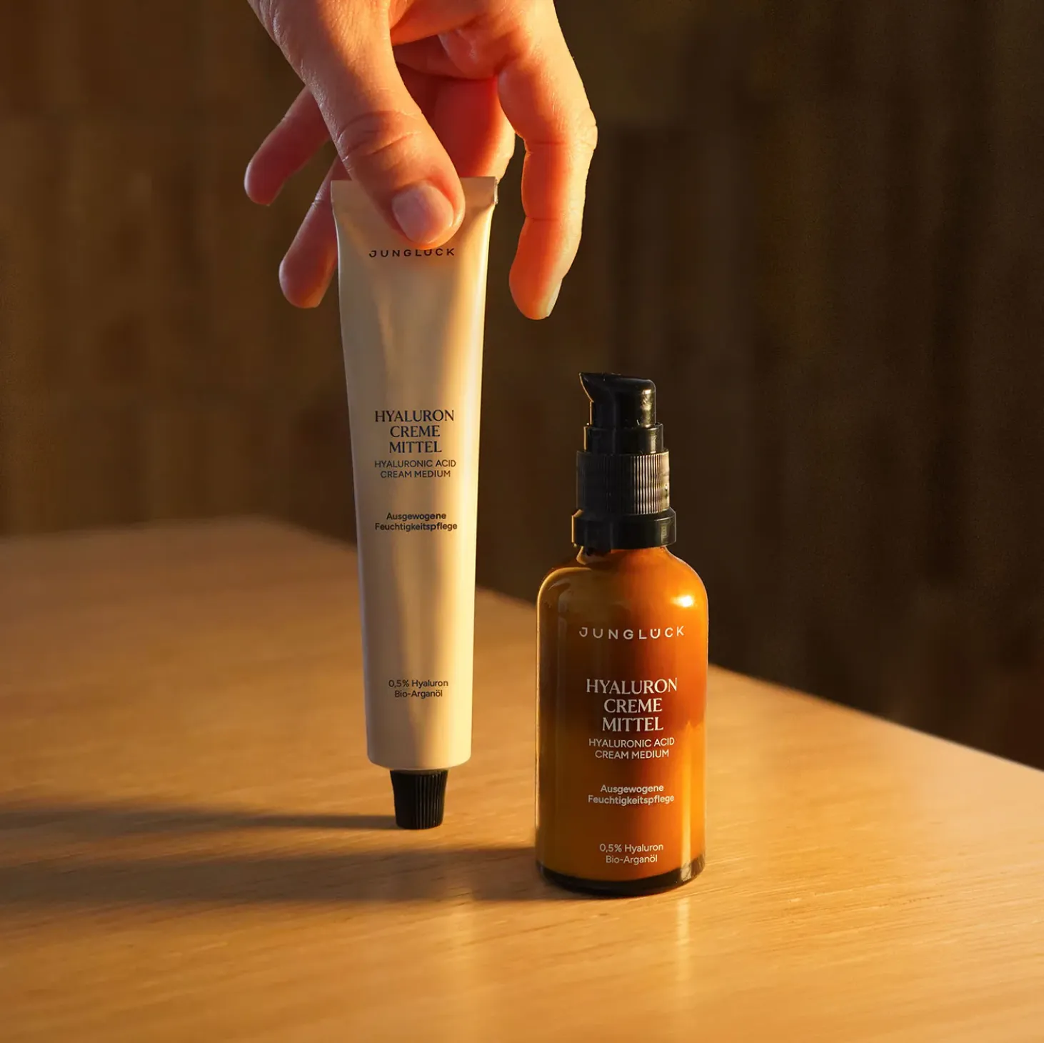 Hand holding a tube of Junglück Hyaluron Creme Mittel next to a brown pump bottle of the same product on a wooden surface.