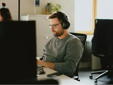 Ein Mann mit Kopfhörern und Brille sitzt in einem Büro an einem Schreibtisch und arbeitet an einem Computer.