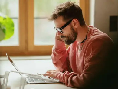 Ein nachdenklich dreinblickender Mann mit Brille und hellrotem Pullover sitzt am Schreibtisch und tippt während eines Telefonats auf seinem Laptop.