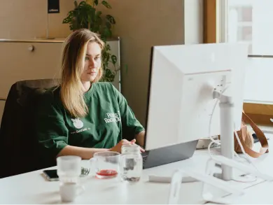 Eine Frau in einem grünen Shirt sitzt in einem gut beleuchteten Raum an einem Schreibtisch mit einem Laptop und einem externen Bildschirm.
