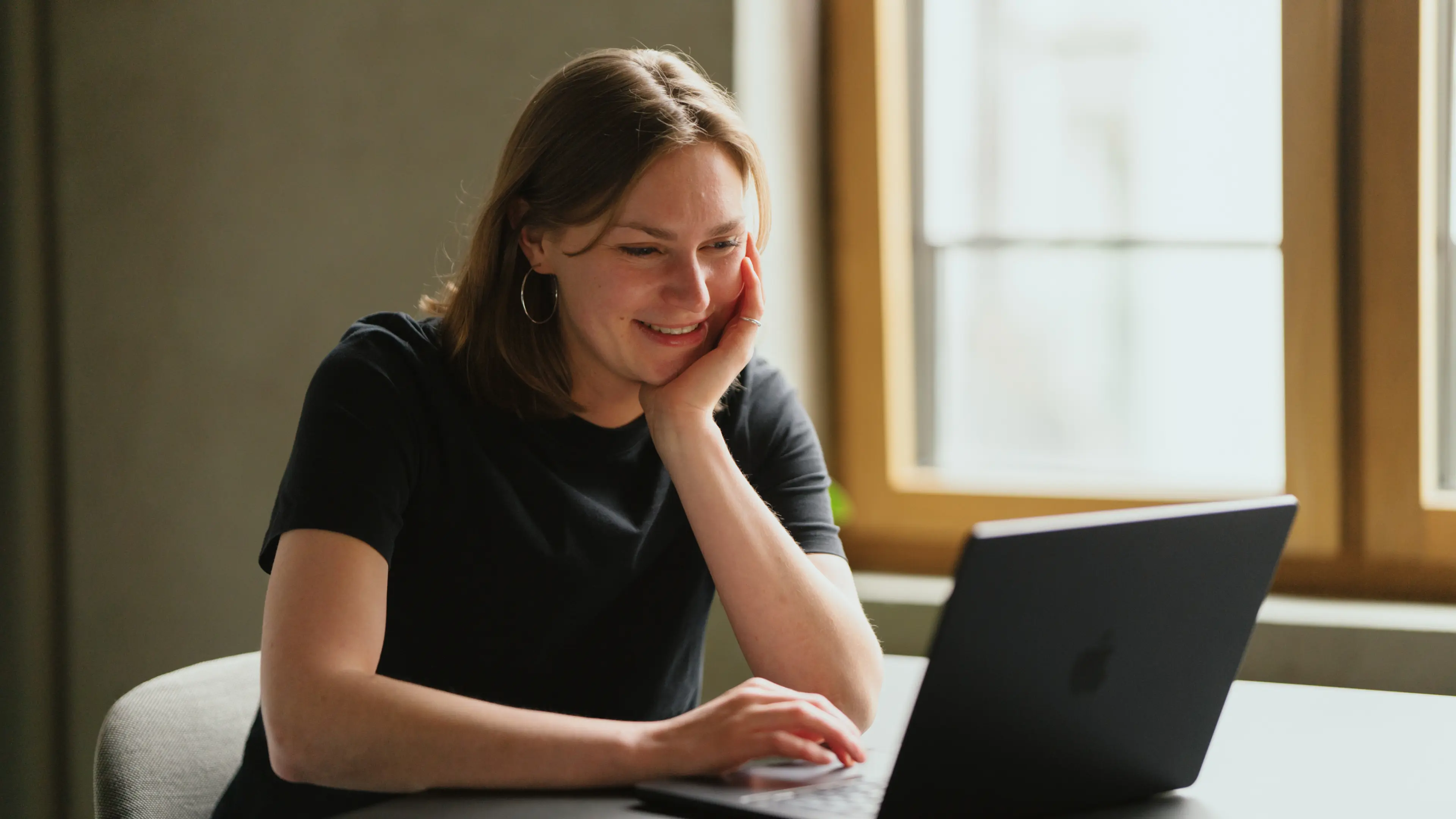 Eine Frau in einem schwarzen Shirt lächelt und tippt auf einem Laptop in der Nähe eines Fensters.