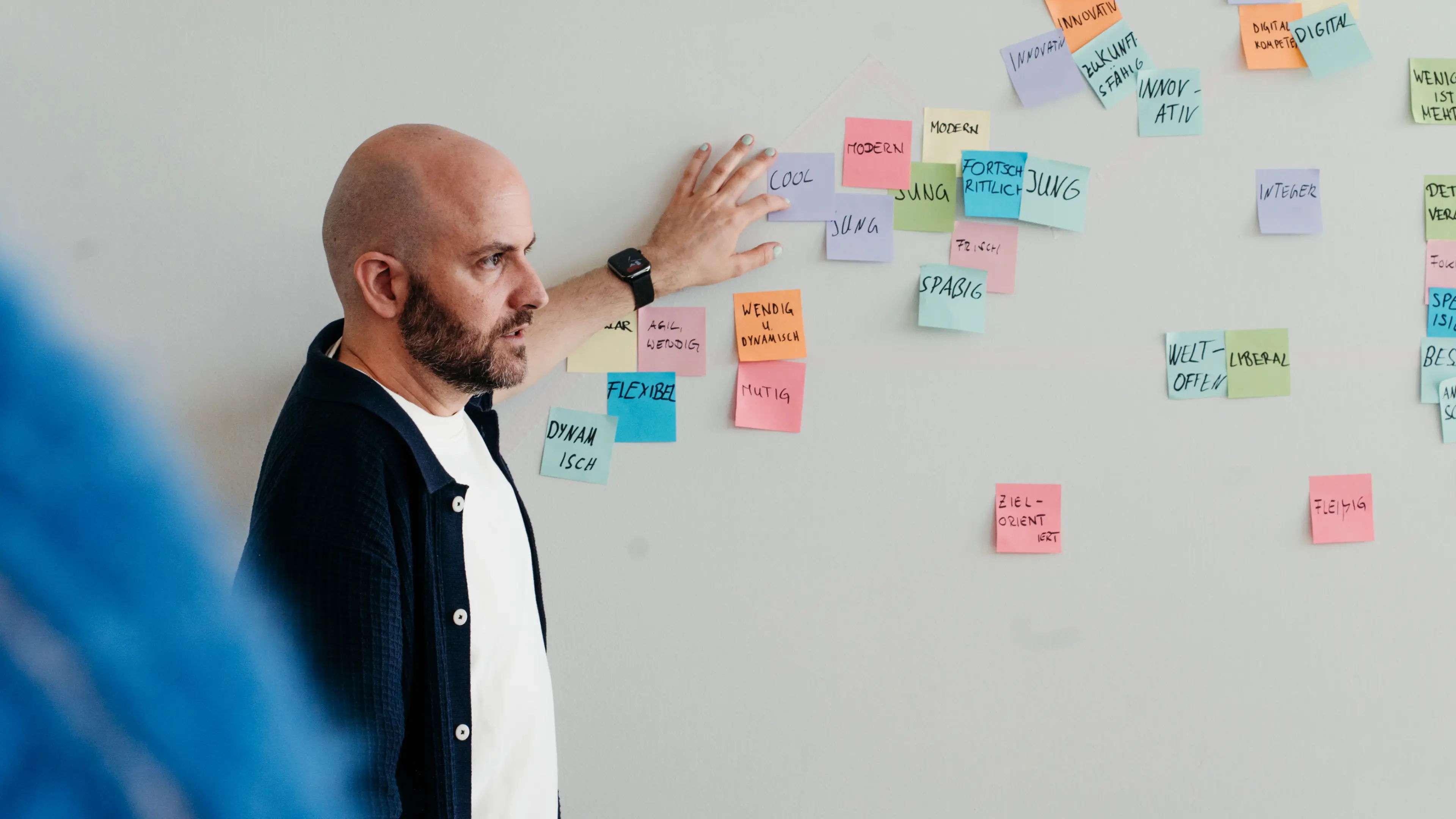 Neben einer mit bunten, beschrifteten Post-its übersäten Wand steht ein glatzköpfiger Mann in einer schwarzen Strickjacke.