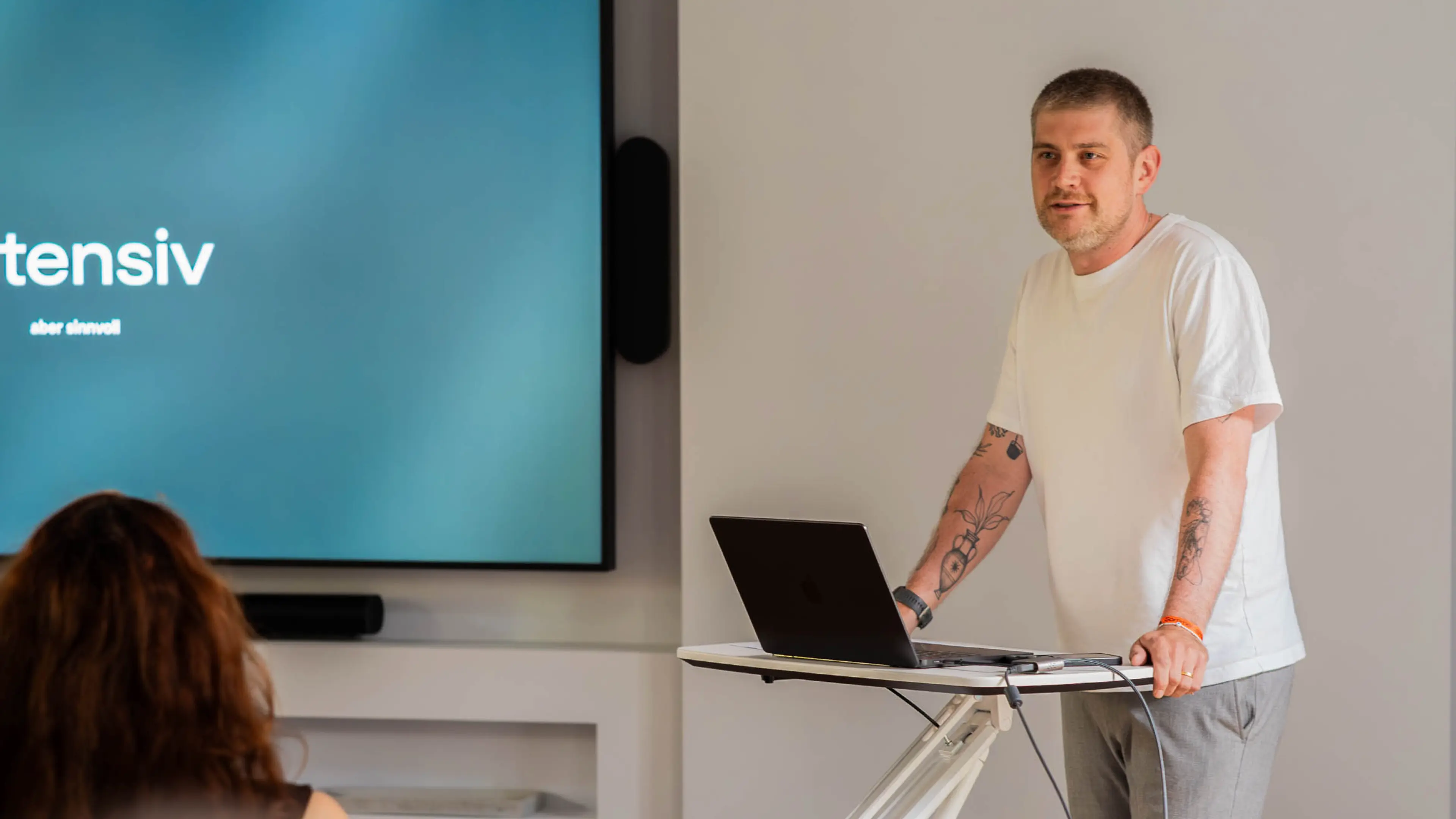 Ein Mann in einem weißen T-Shirt, der vor einem Laptop und neben einem Bildschirm steht und eine Präsentation hält.