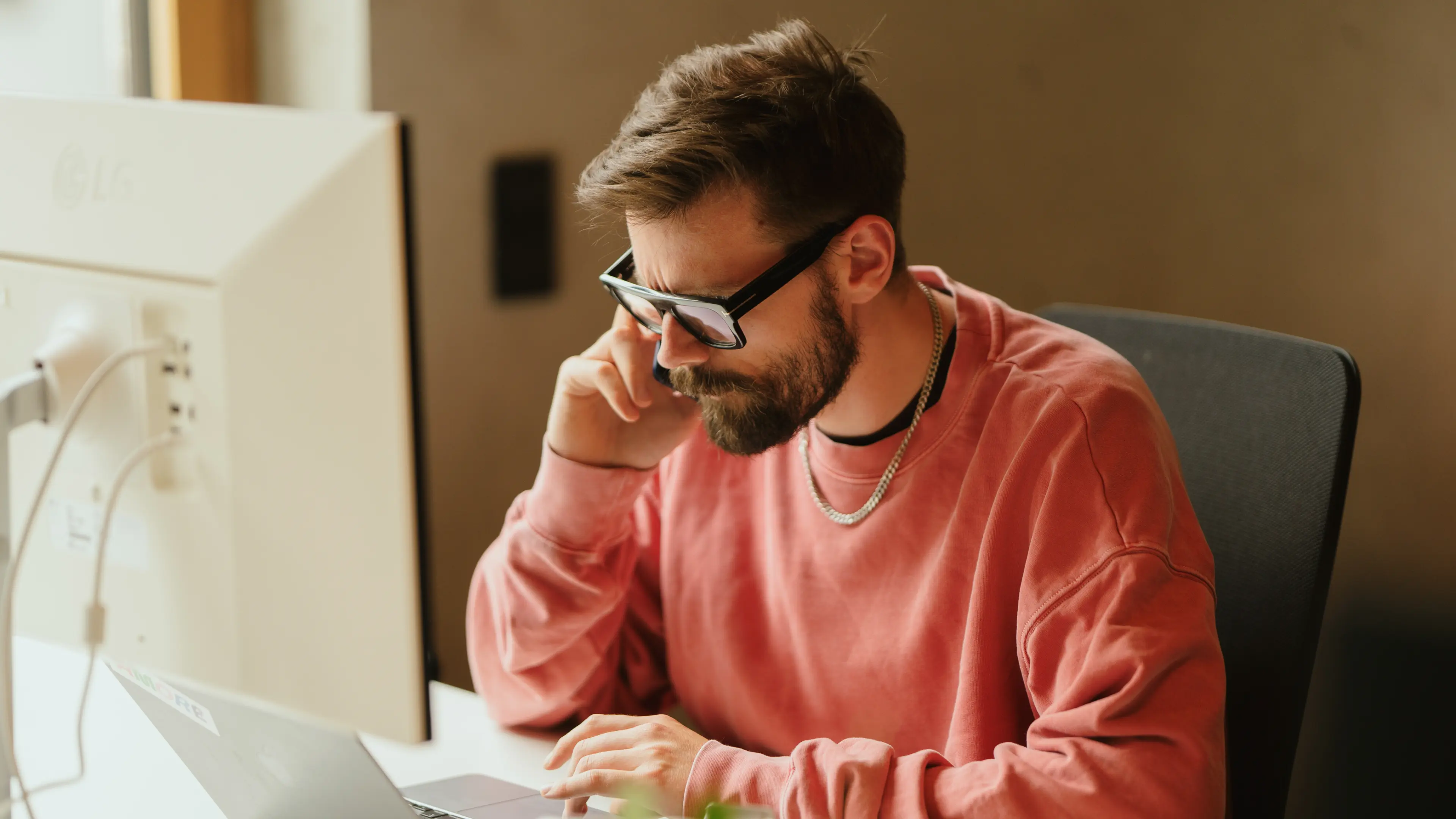 Ein nachdenklich dreinblickender Mann mit Brille und hellrotem Pullover sitzt am Schreibtisch und tippt während eines Telefonats auf seinem Laptop.