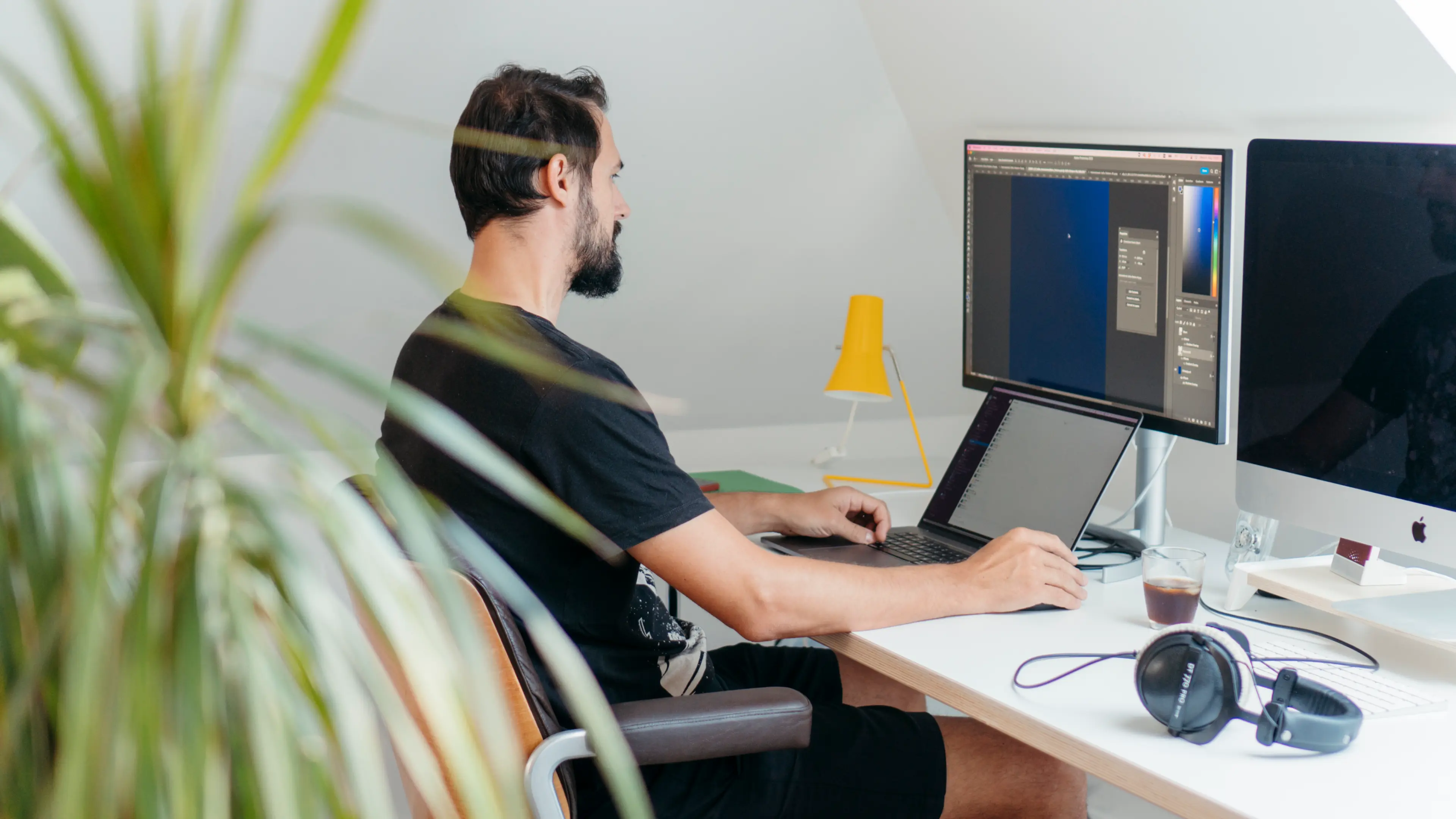 Ein Mann arbeitet an einem Laptop und einem externen Monitor in einem gut beleuchteten Raum mit einer Pflanze.