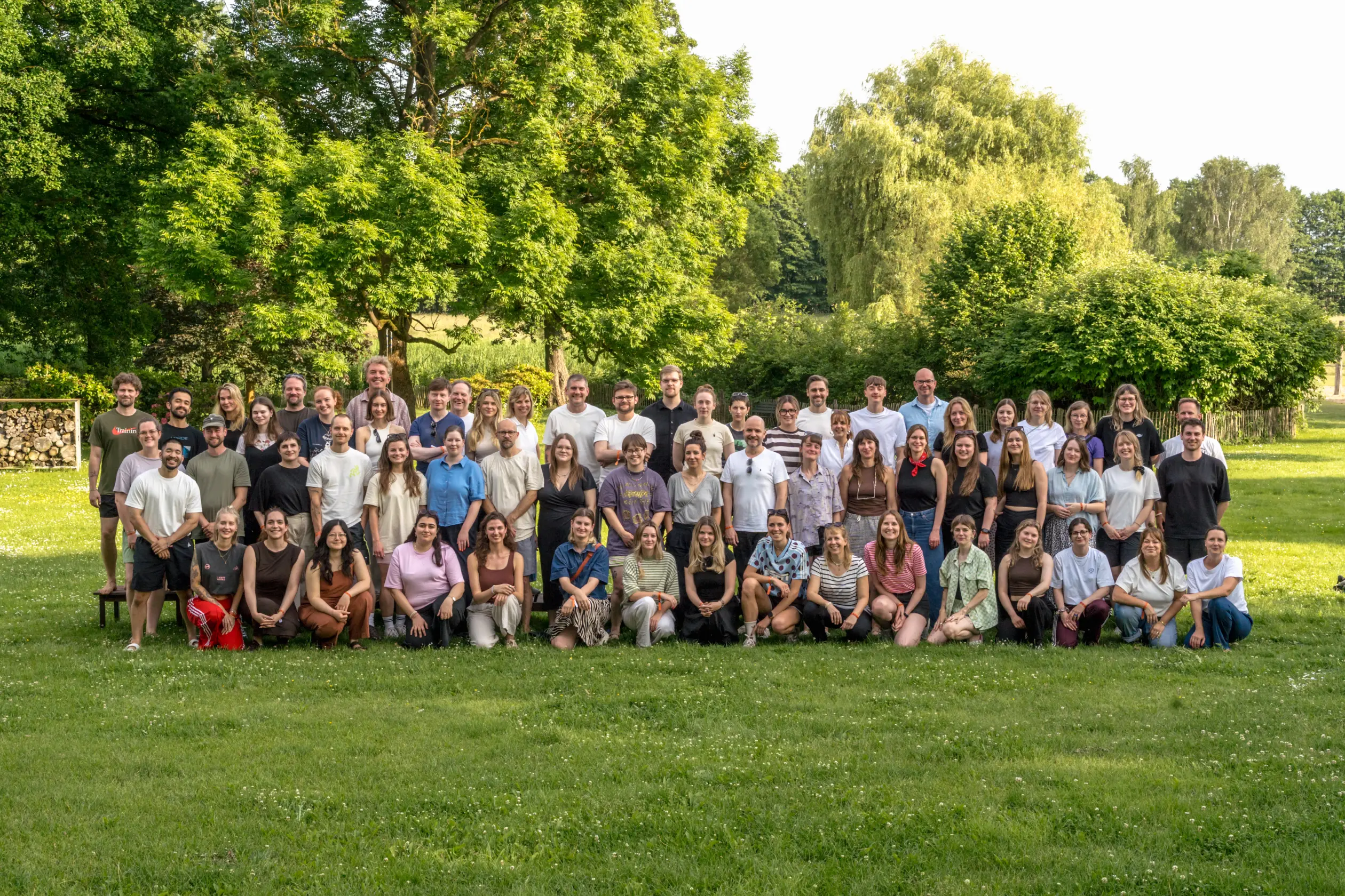 Eine große Menschengruppe posiert für ein Foto auf einer Wiese mit Bäumen im Hintergrund.
