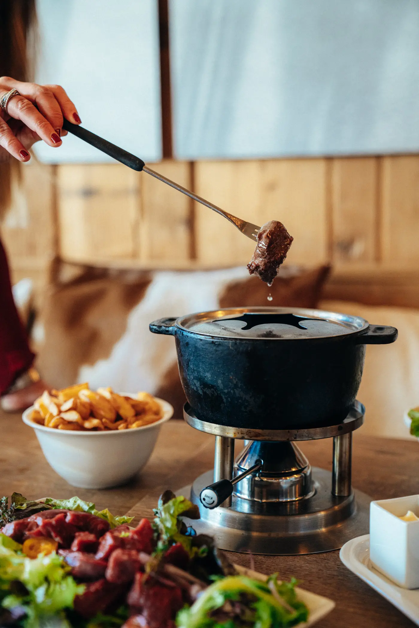 Hand holding fork with cooked meat dripping over a tabletop fondue pot with sides of chips and salad.