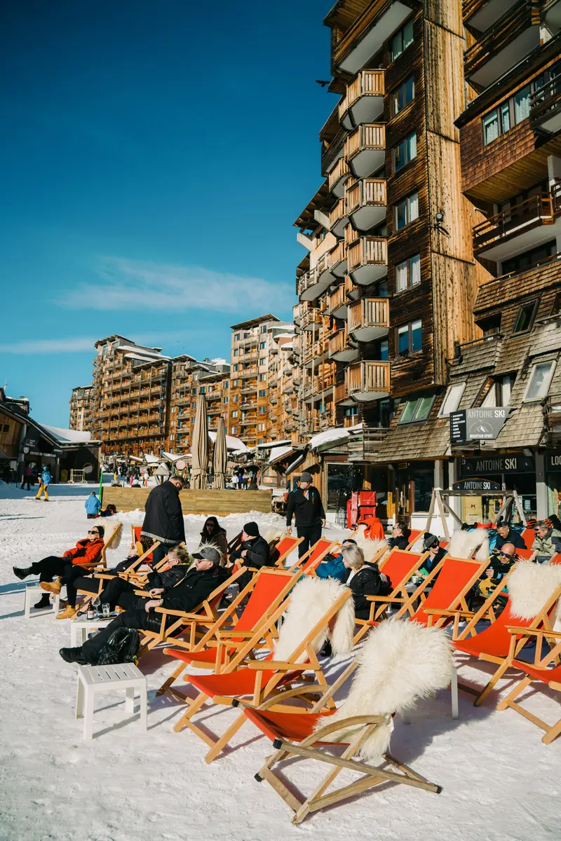 Des personnes se détendent sur des chaises longues orange, dans la neige, près de chalets de ski en bois et sous un ciel bleu limpide, restaurant La Brasserie à Avoriaz, Haute-Savoie, France.