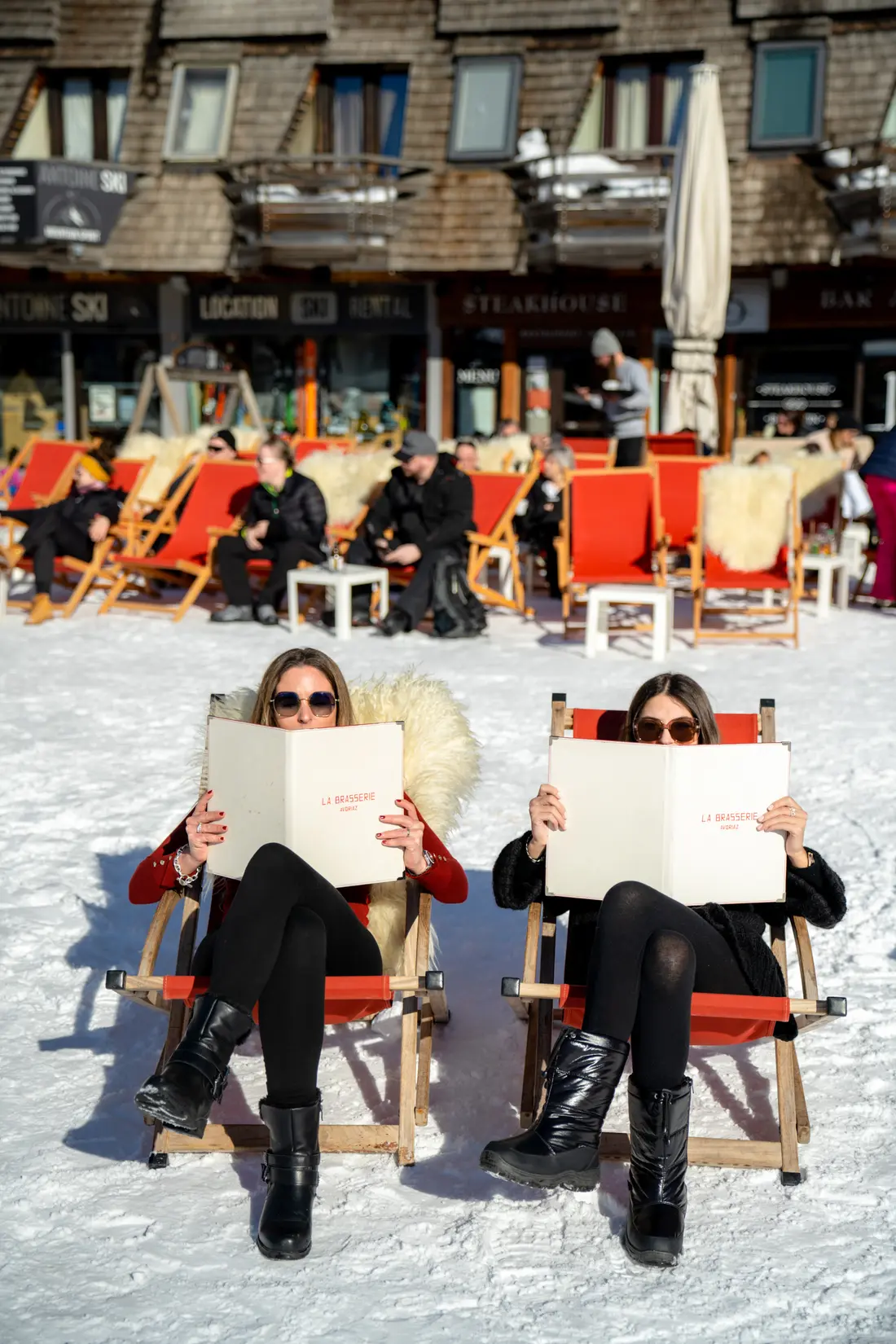 Deux femmes portant des lunettes de soleil, assises sur des chaises rouges dans la neige, lisent les menus de La Brasserie, restaurant La Brasserie à Avoriaz, Haute-Savoie, France.