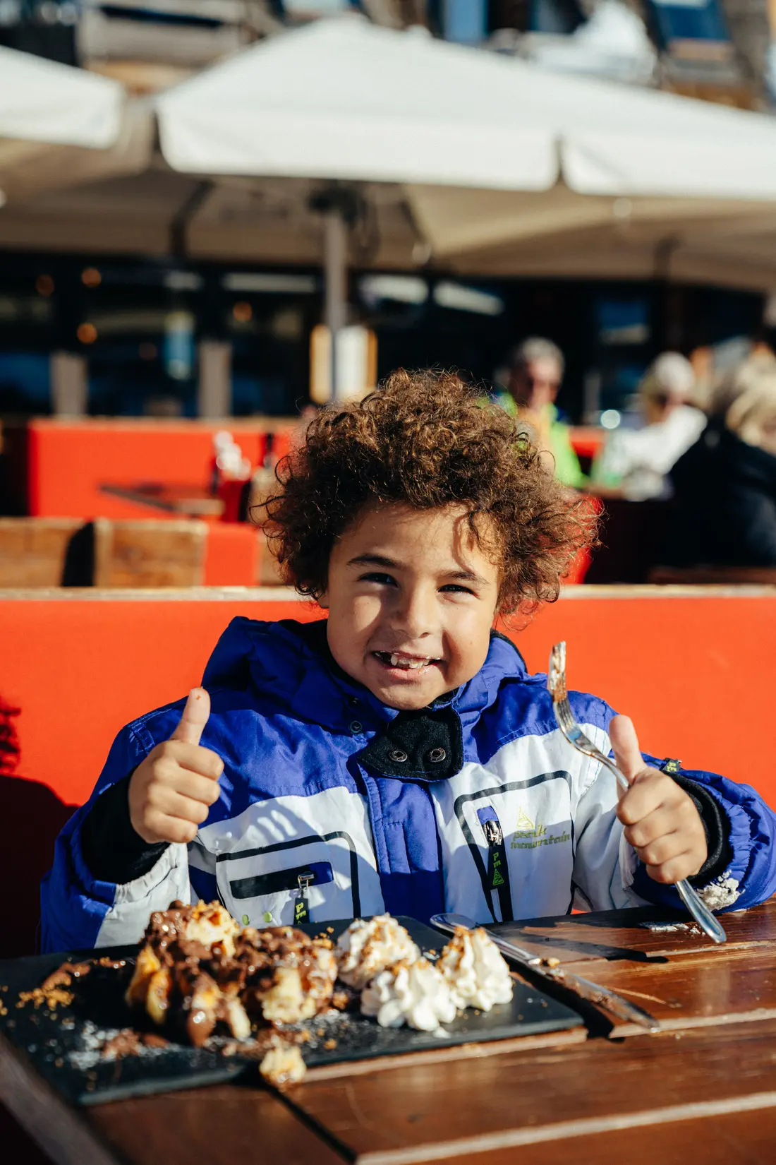 Un enfant en veste bleue sourit et fait un signe du pouce levé, avec un dessert sur la table devant lui, restaurant La Brasserie à Avoriaz, Haute-Savoie, France.