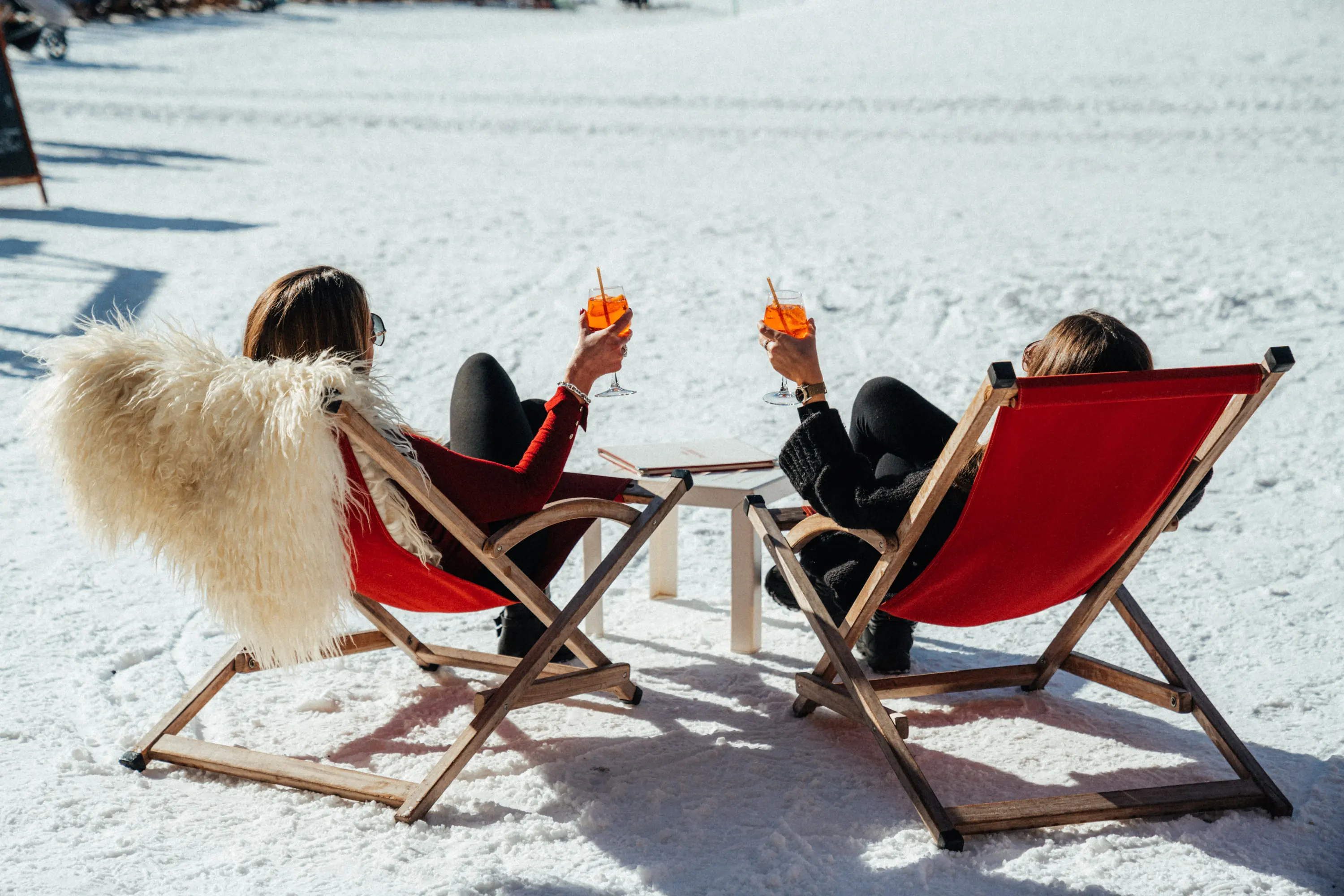 Deux femmes assises dans des chaises longues rouges sur la neige, entrechoquant leurs verres contenant des boissons orange, restaurant La Brasserie à Avoriaz, Haute-Savoie, France.