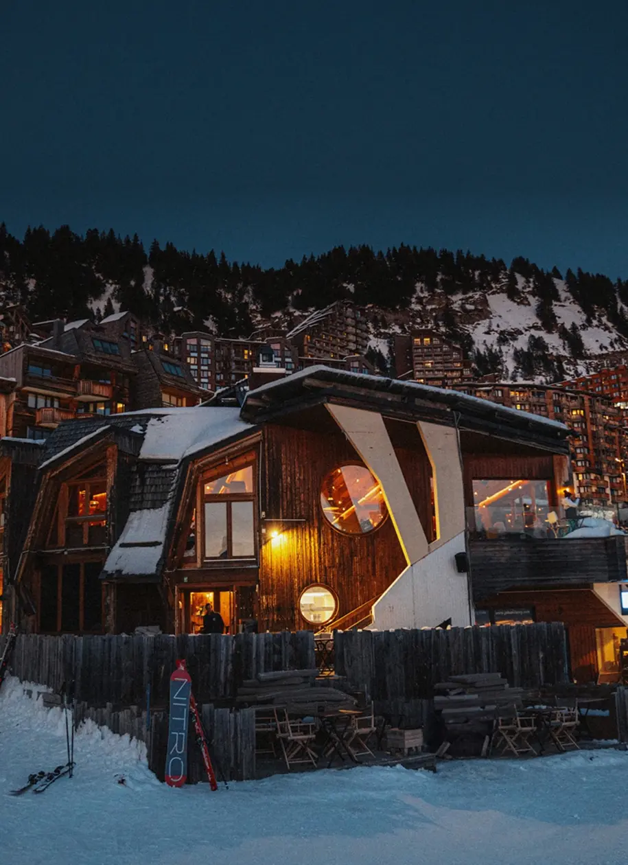Chalet de ski en bois enneigé, chaleureusement éclairé au crépuscule, avec des bâtiments de montagne et des pins en arrière-plan, restaurant La Brasserie à Avoriaz, Haute-Savoie, France.
