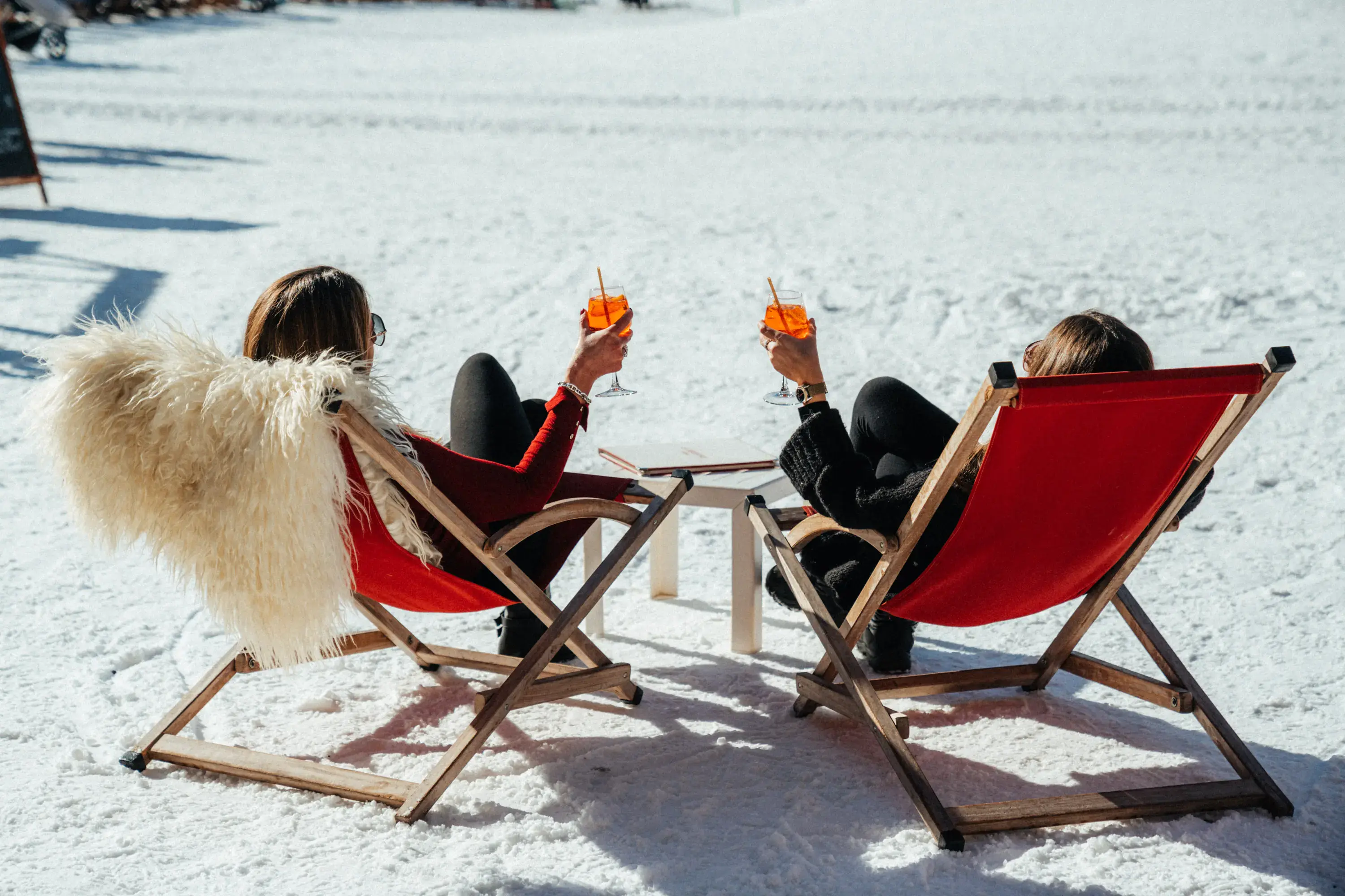 Deux femmes assises sur des chaises longues rouges dans la neige, trinquant avec des boissons à l'orange par une journée ensoleillée, restaurant La Brasserie à Avoriaz, Haute-Savoie, France.
